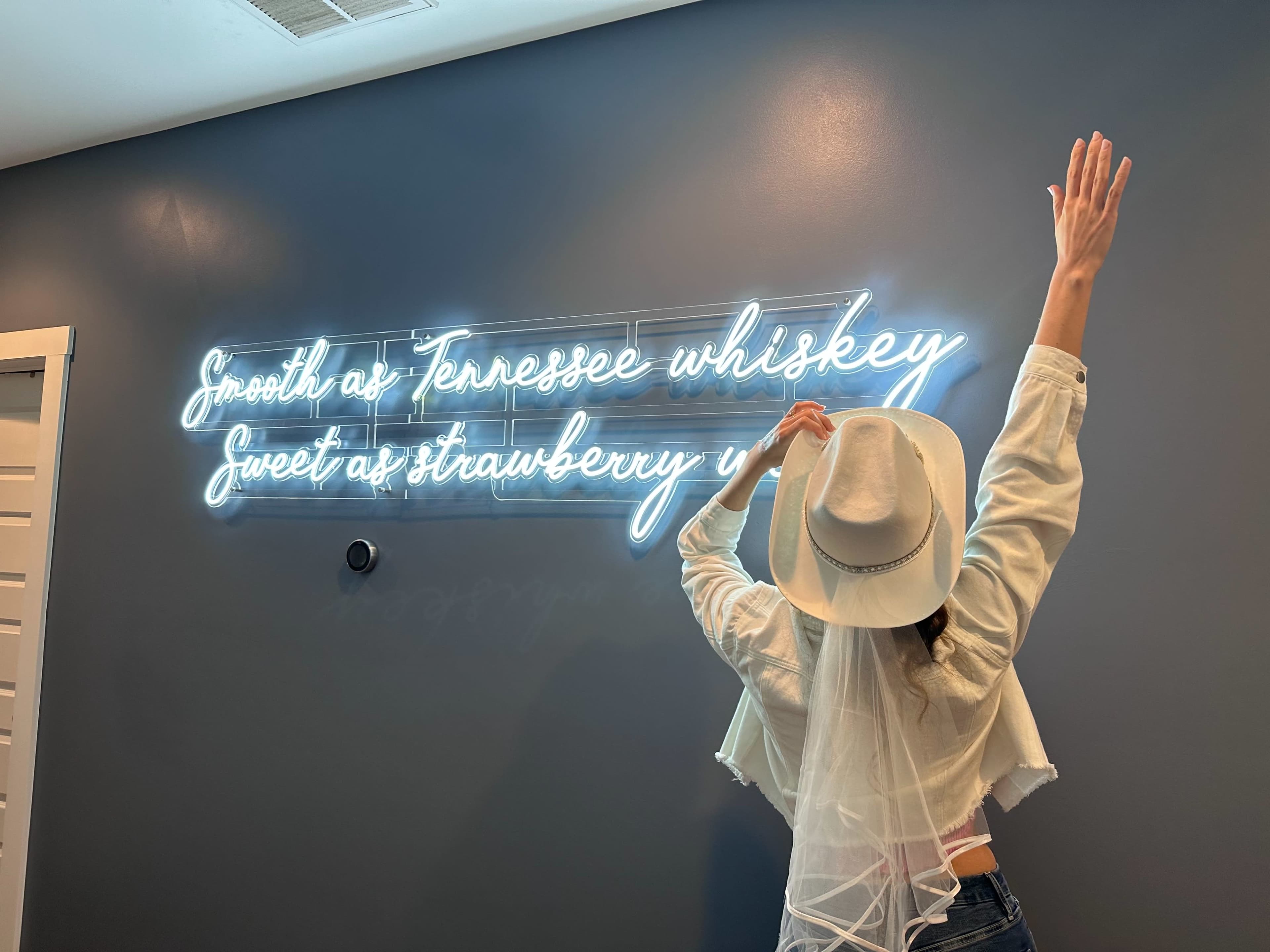 A person with raised arms stands in front of a neon sign that reads "Smooth as Tennessee whiskey, Sweet as strawberry wine" against a blue wall.