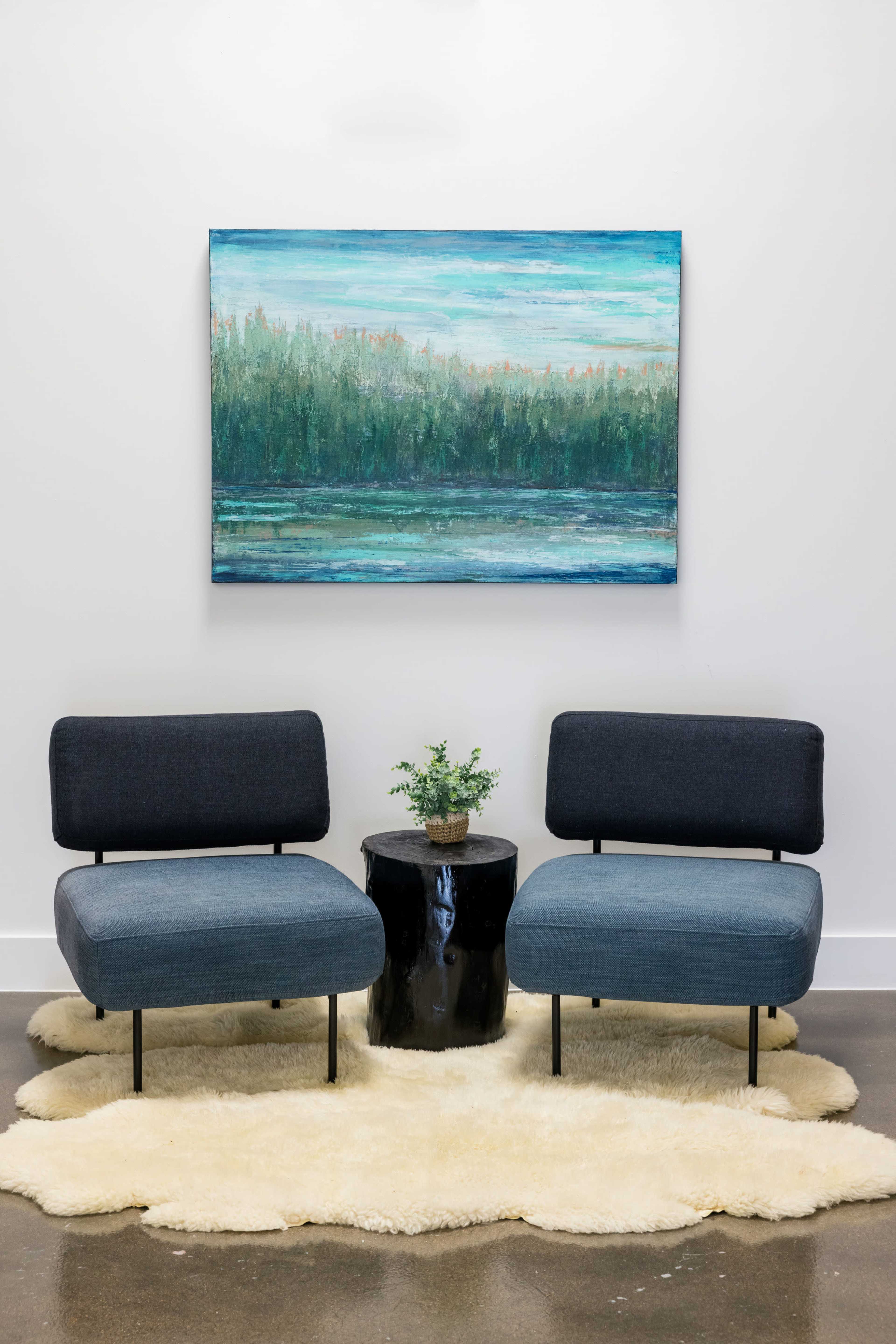 Two modern chairs are positioned facing each other on a faux fur rug, with a small plant and a black side table between them, under a landscape painting.