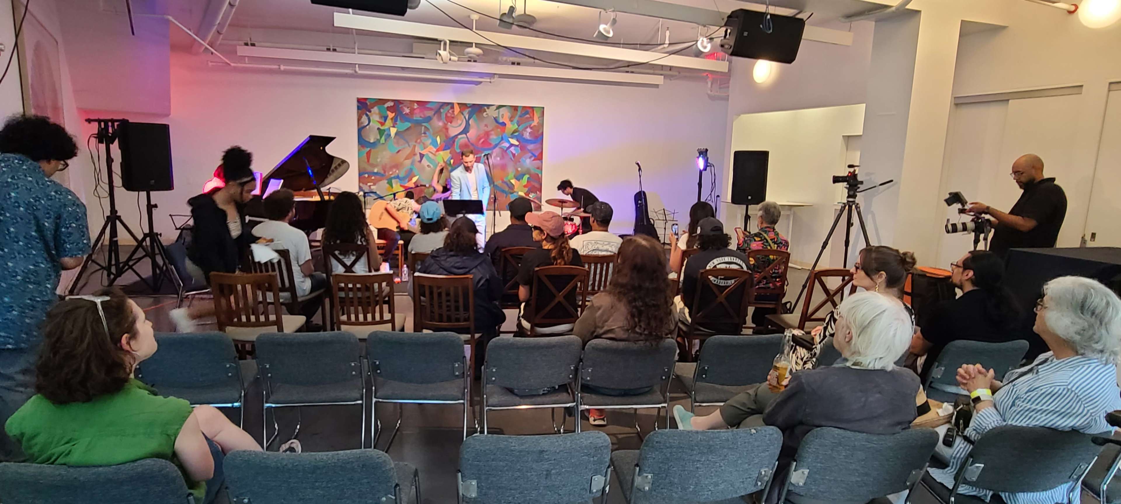 A diverse group of people is seated in a gallery space facing a stage with performers, while cameras and lighting equipment are set up for a live event.
