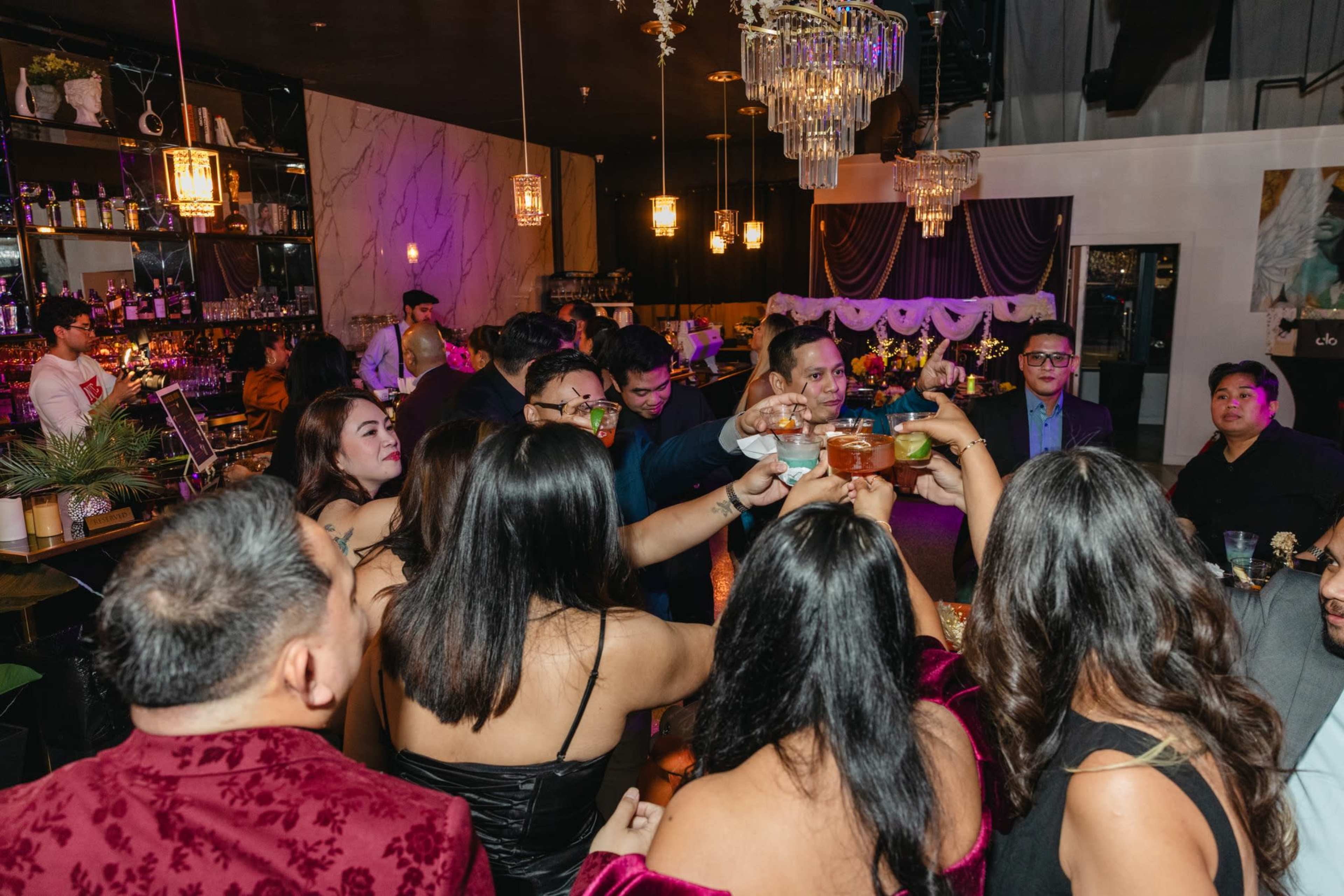 A group of people is raising their drinks in a toast at a festive gathering in a stylish bar setting.