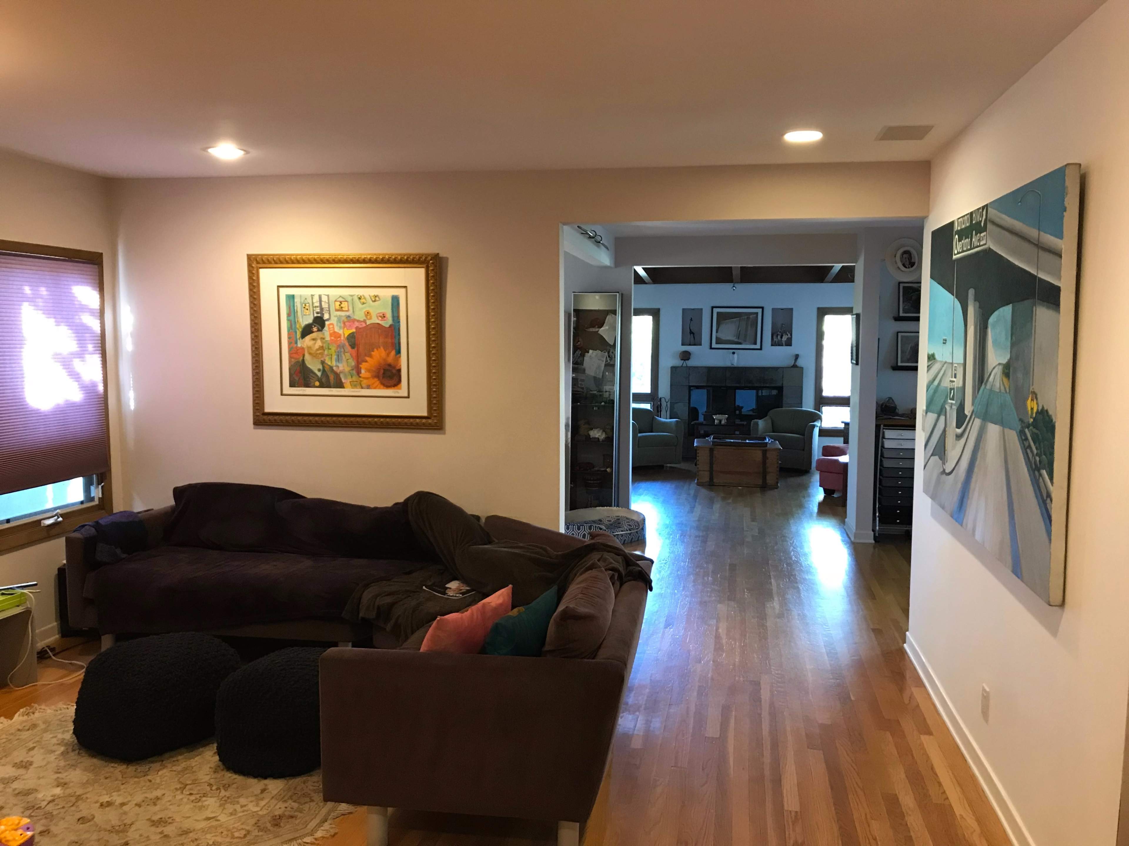 A living room features a dark couch, a framed painting on the wall, and a view of another room with wooden flooring and furniture.