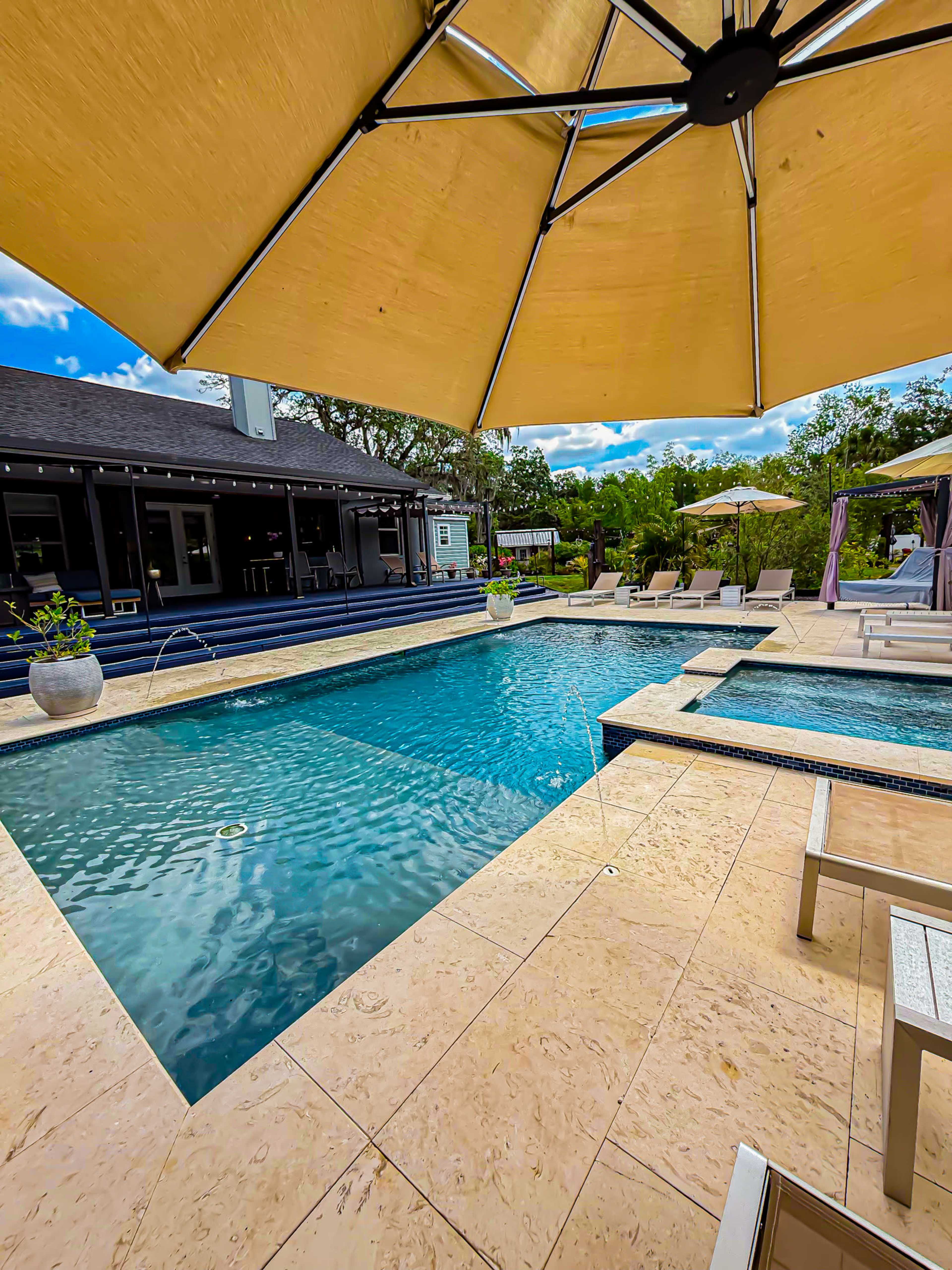 A patio area with a swimming pool is shaded by a large umbrella, surrounded by lounge chairs and greenery.