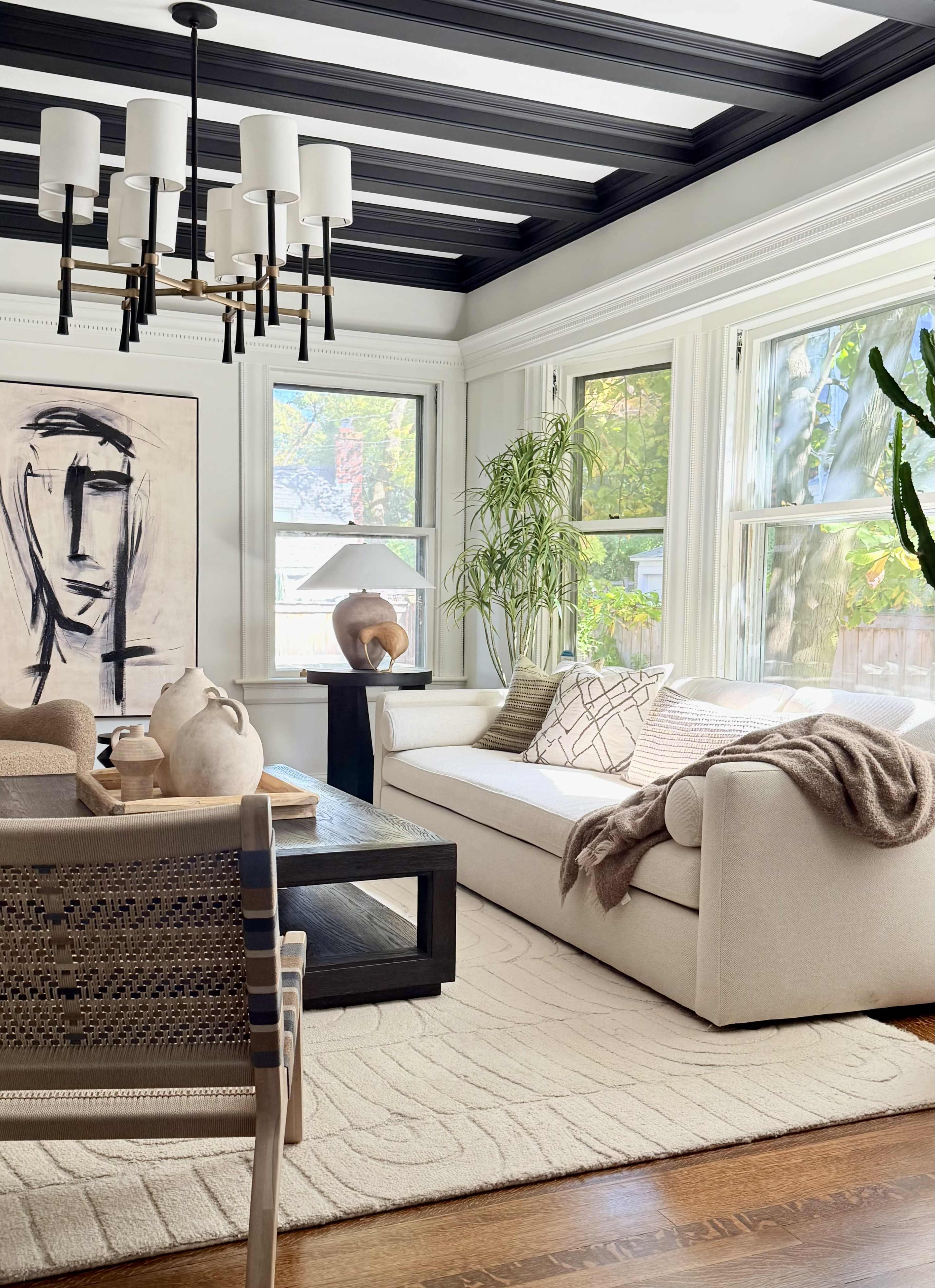 The image shows a well-lit living room featuring a light-colored sofa, large windows, a coffee table with decorative objects, and a mix of plants and artwork.