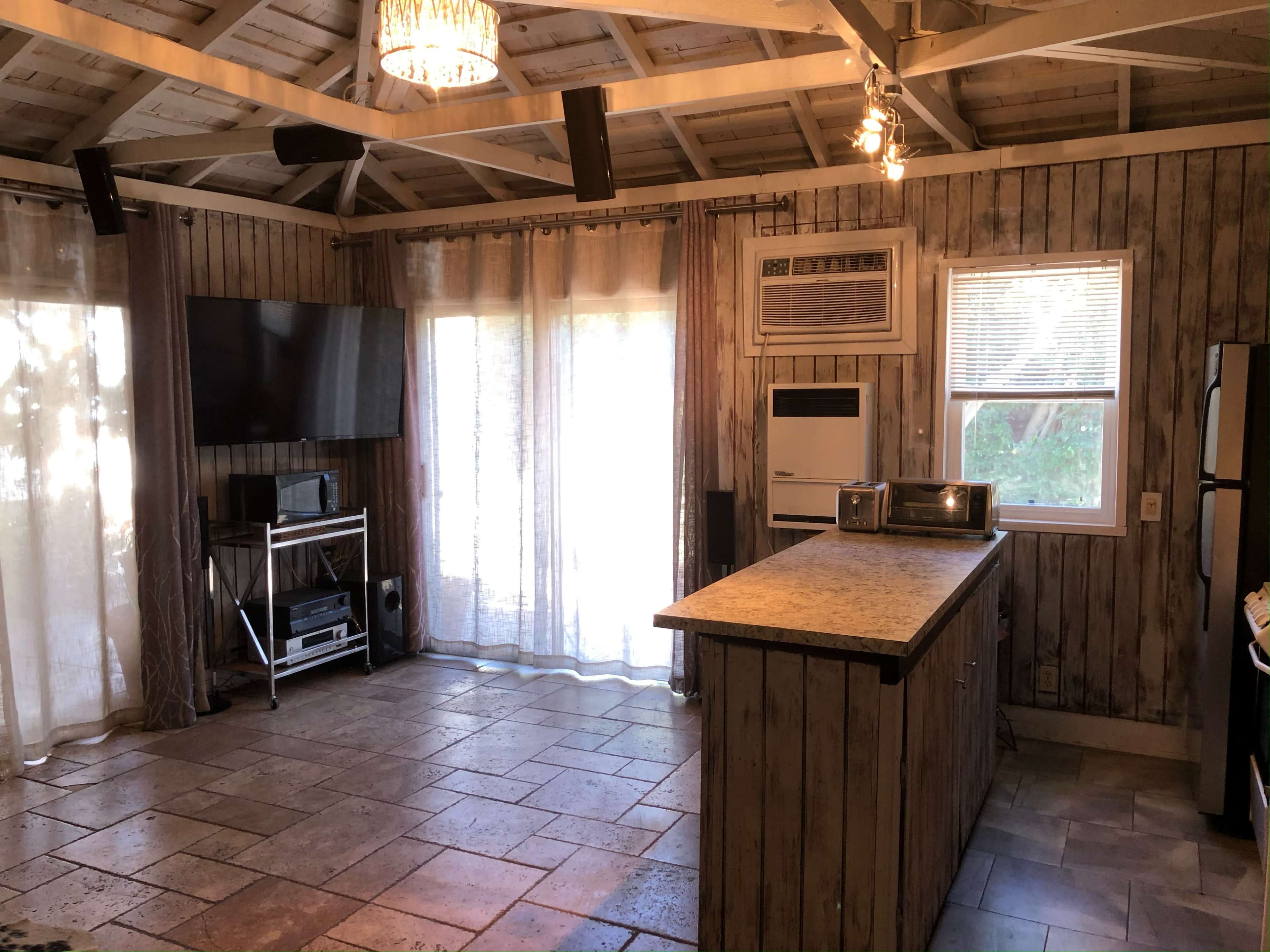 The image shows a rustic interior space with a kitchen island, a television mounted on a wall, and large windows covered by sheer curtains.
