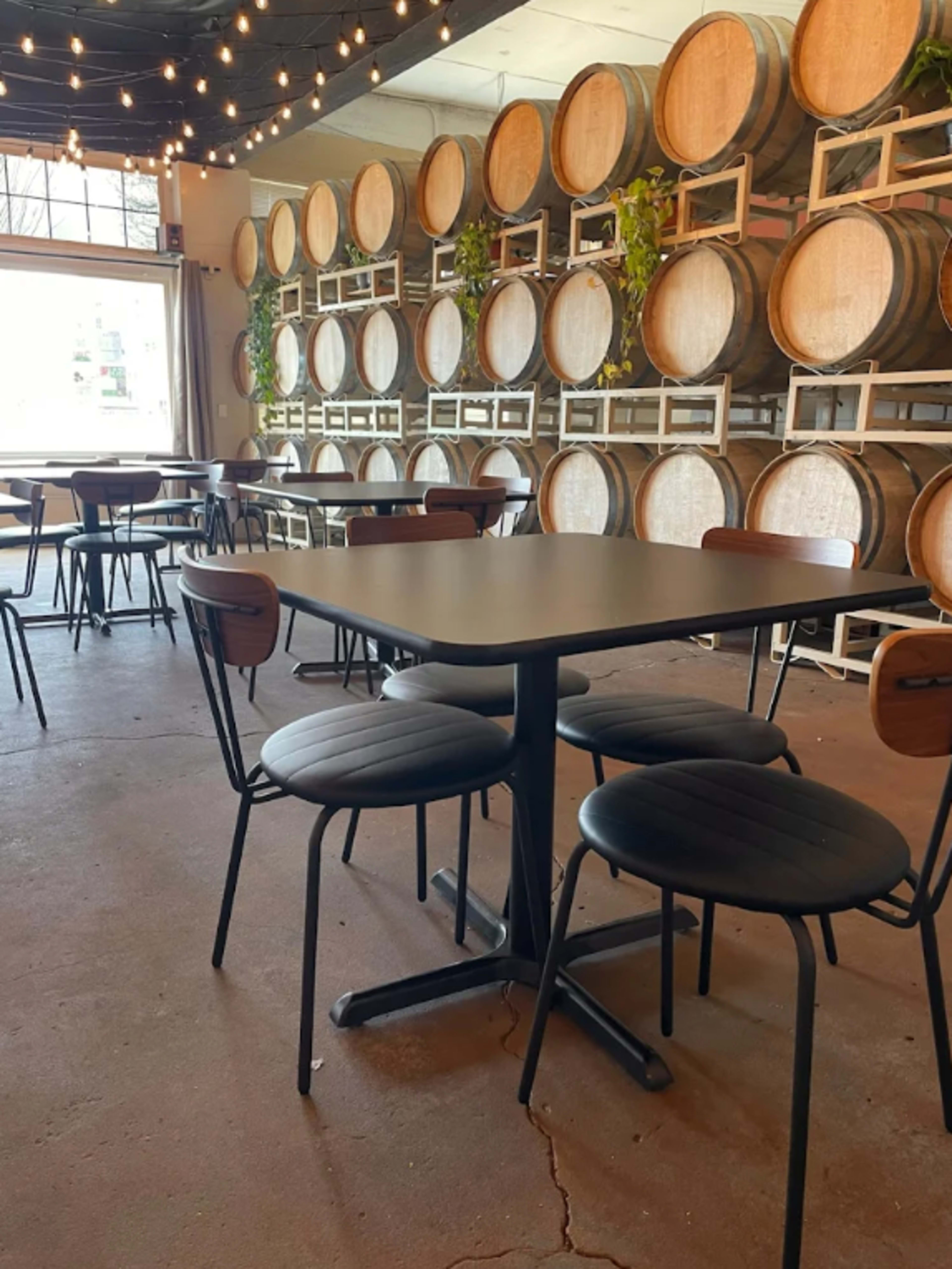 The image shows a dining area with a black table and metal chairs, set against a wall of wine barrels and string lights.