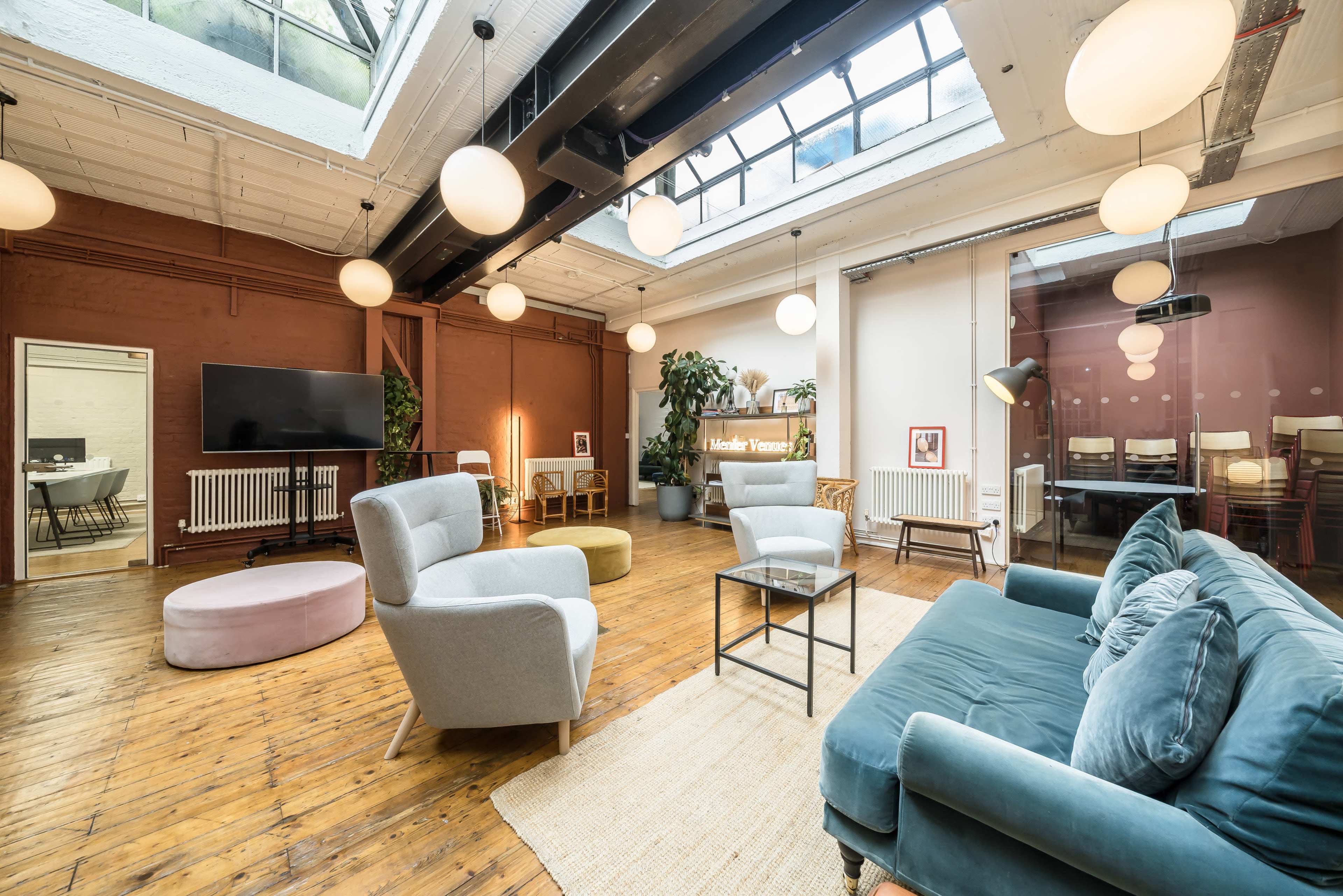 The image depicts a modern, well-lit lounge area with a wooden floor, featuring plush seating arrangements, a low coffee table, and decorative plants.