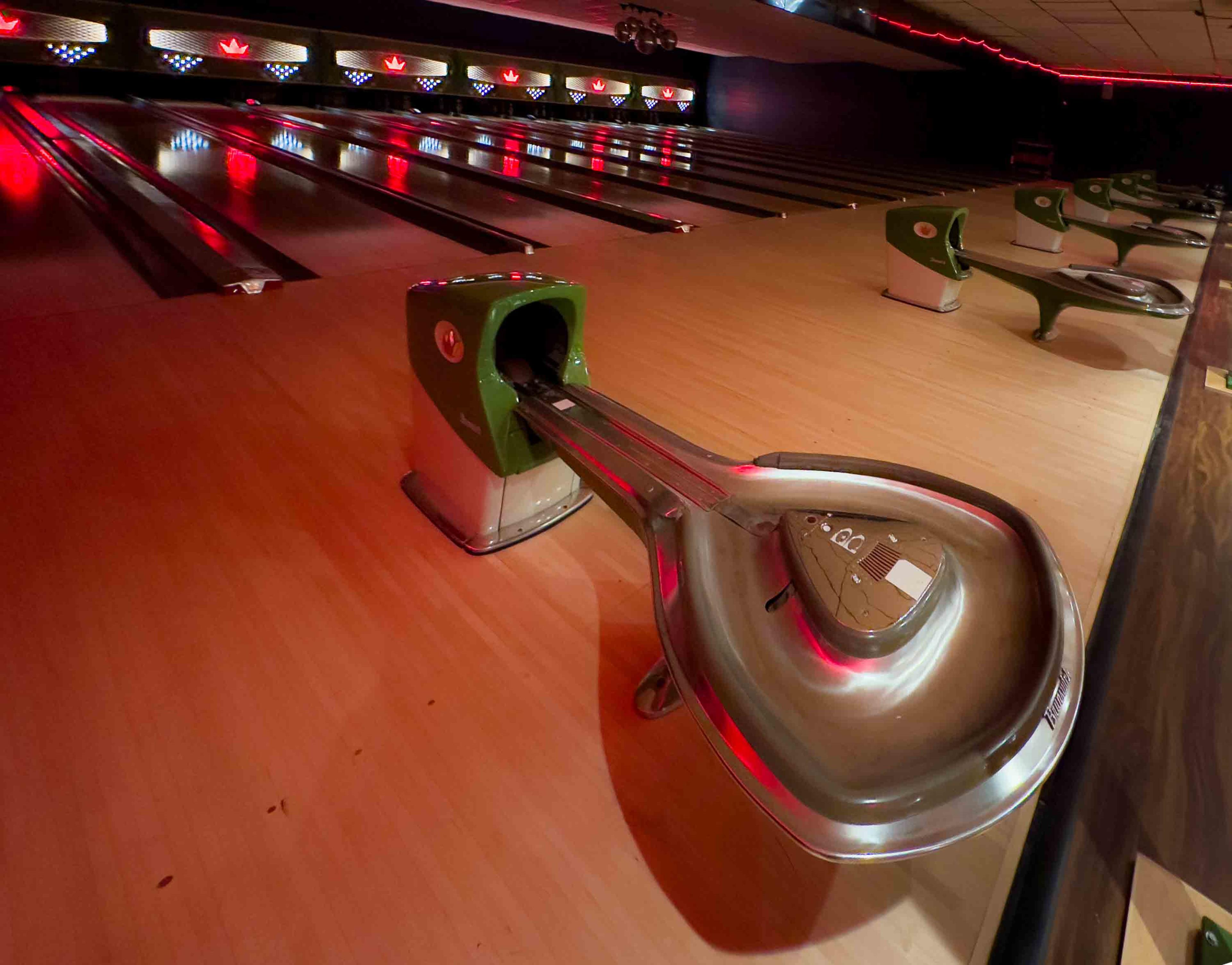 The image shows a bowling alley lane with a metallic ball return system and green seating areas visible in the background.