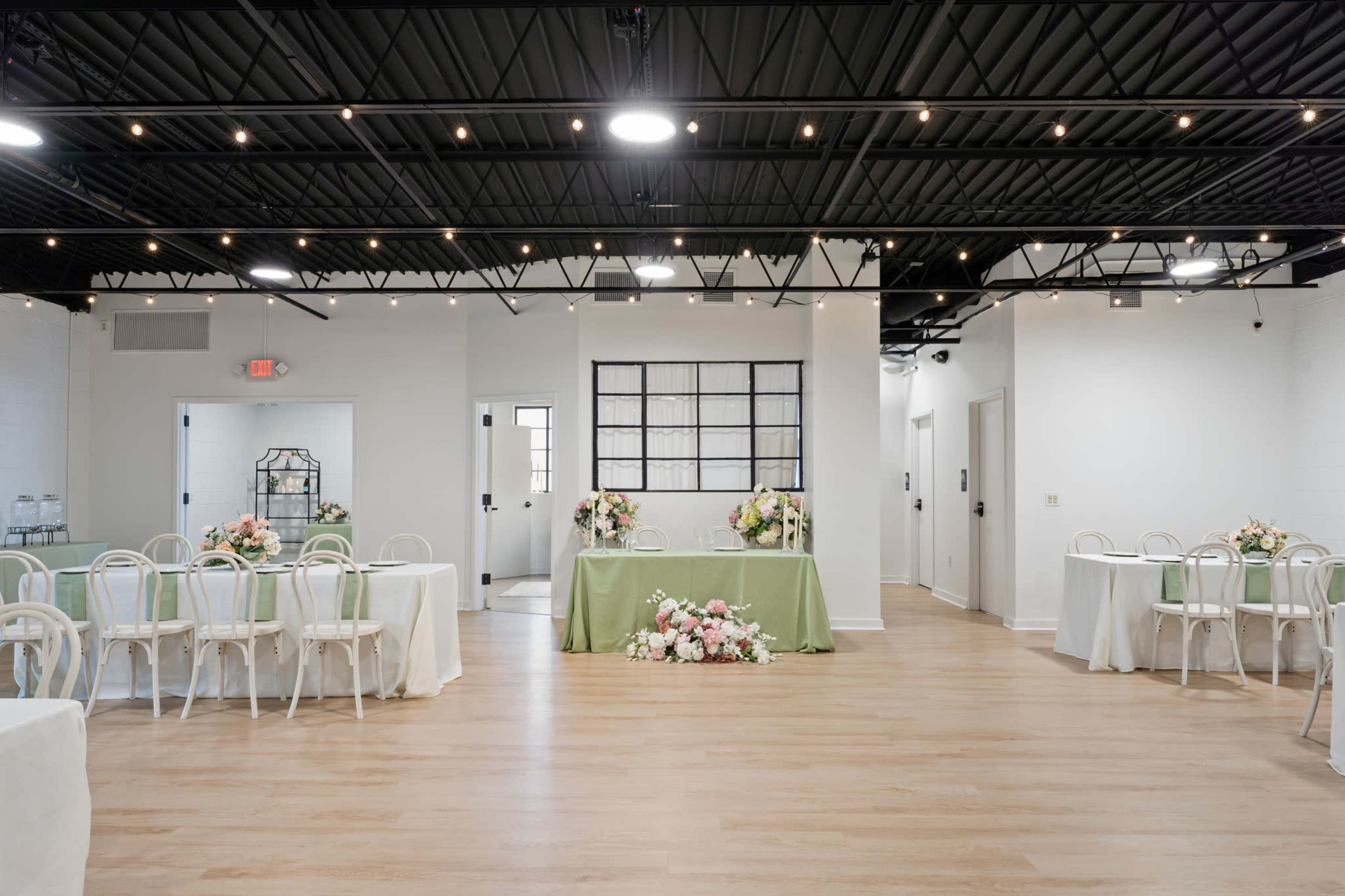The image shows an elegantly arranged event space featuring tables with white chairs and a decorated centerpiece on a green table against a backdrop of string lights and large windows.