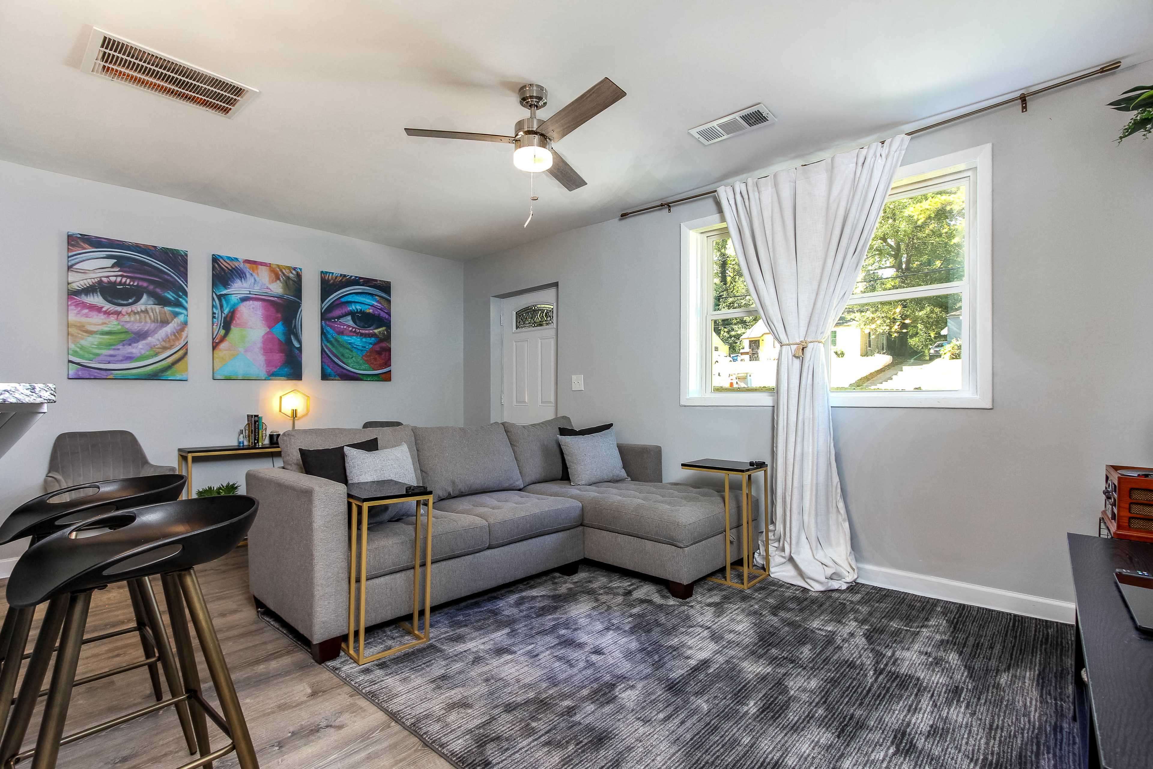 The living room features a gray sectional sofa, a ceiling fan, and colorful framed artwork on the wall, with a window allowing natural light to enter.
