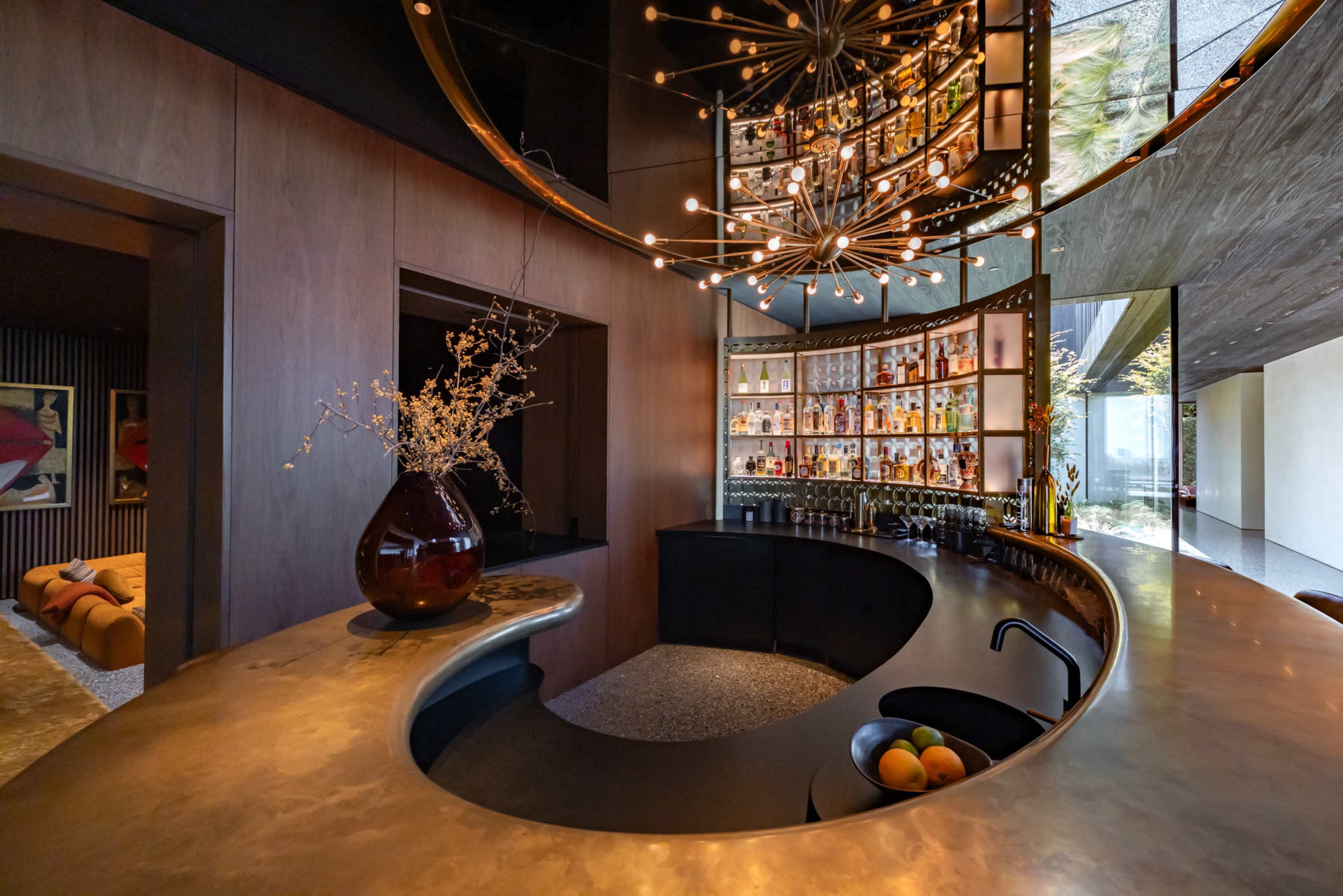 The image shows a modern bar area with a curved countertop, a decorative light fixture, and shelves displaying various bottles.