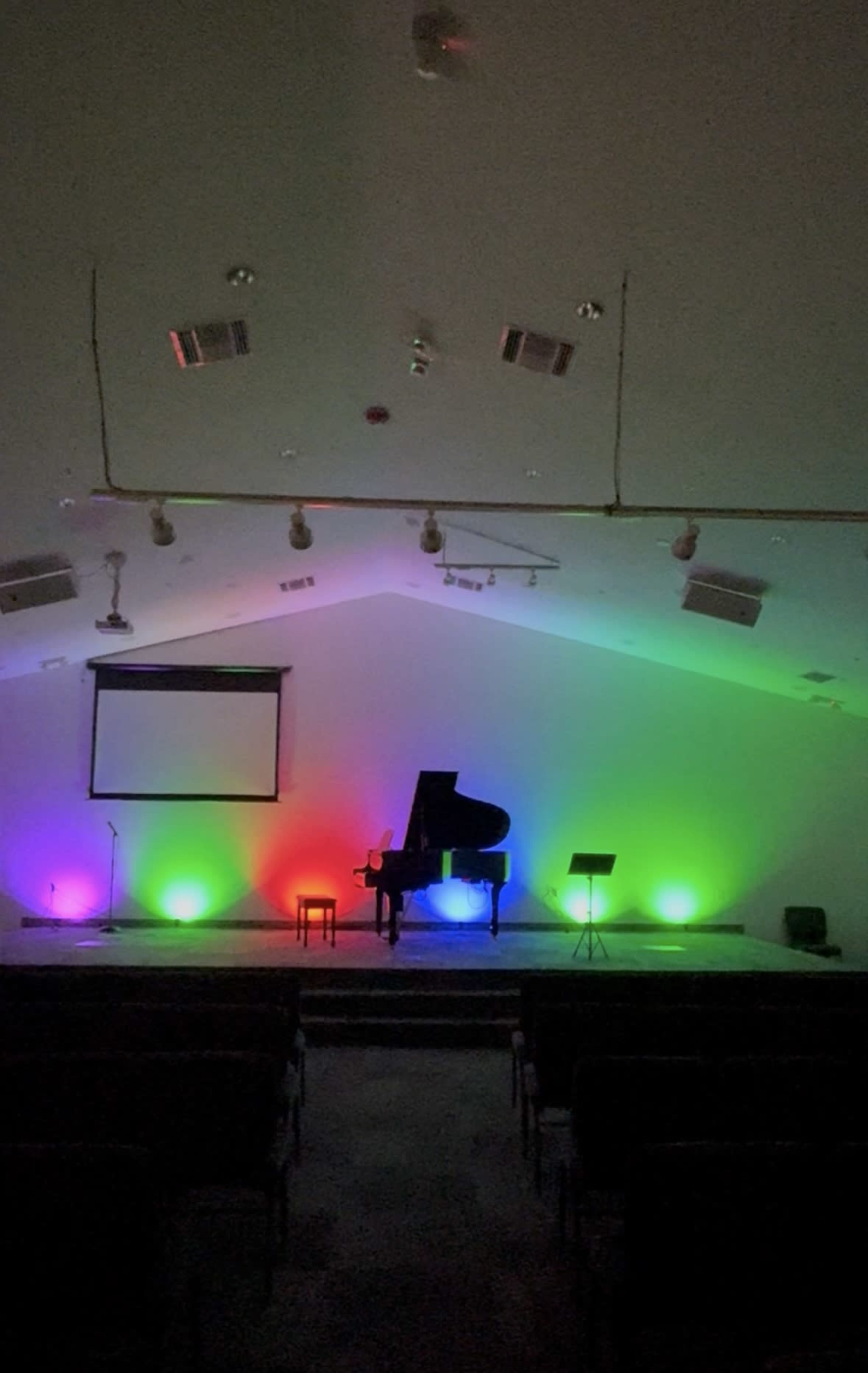 A darkened performance space features a grand piano and a music stand illuminated by colorful stage lights.