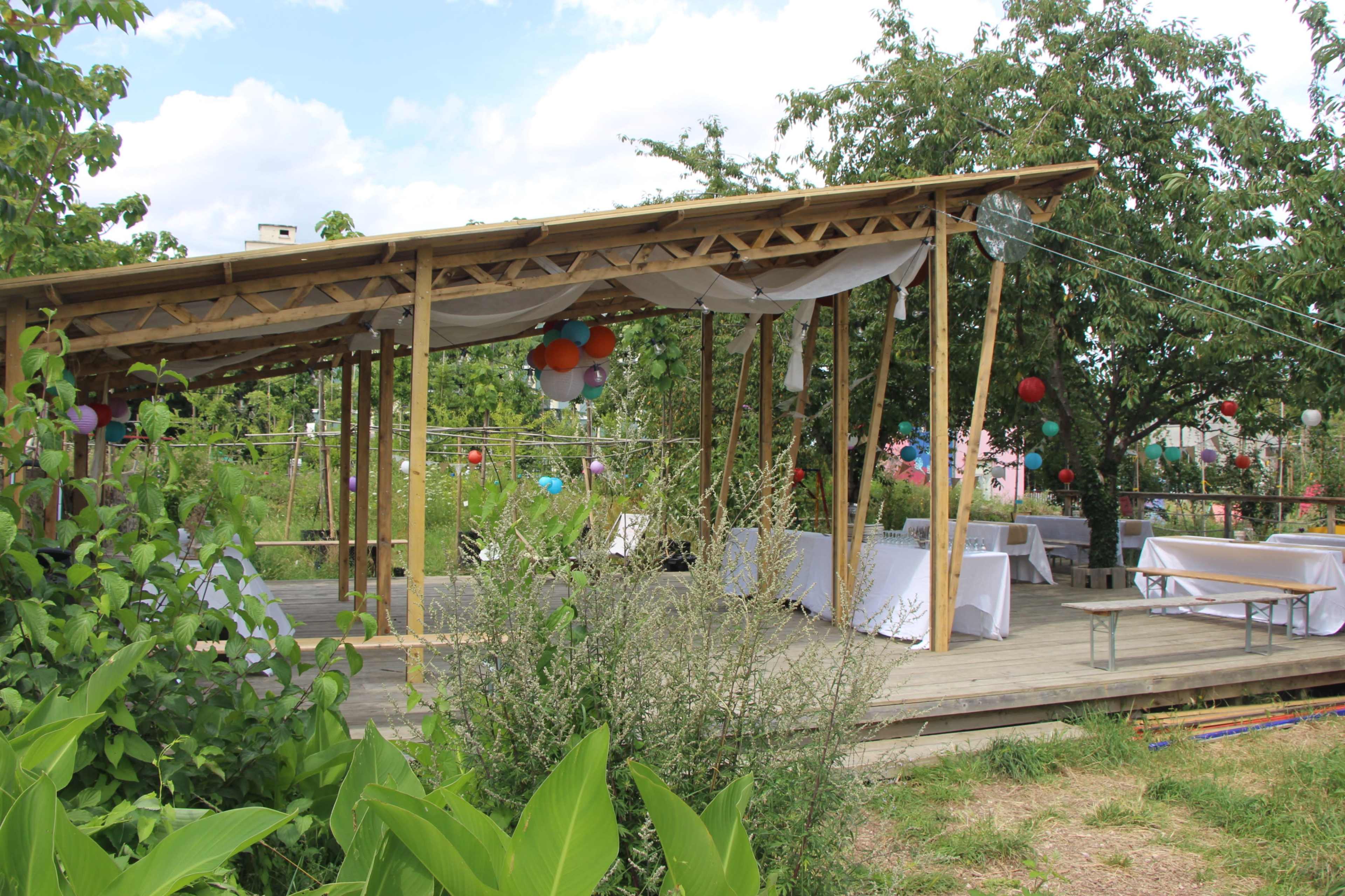 A wooden pergola with draped fabric and tables is set up in a garden, surrounded by greenery and colorful decorations.