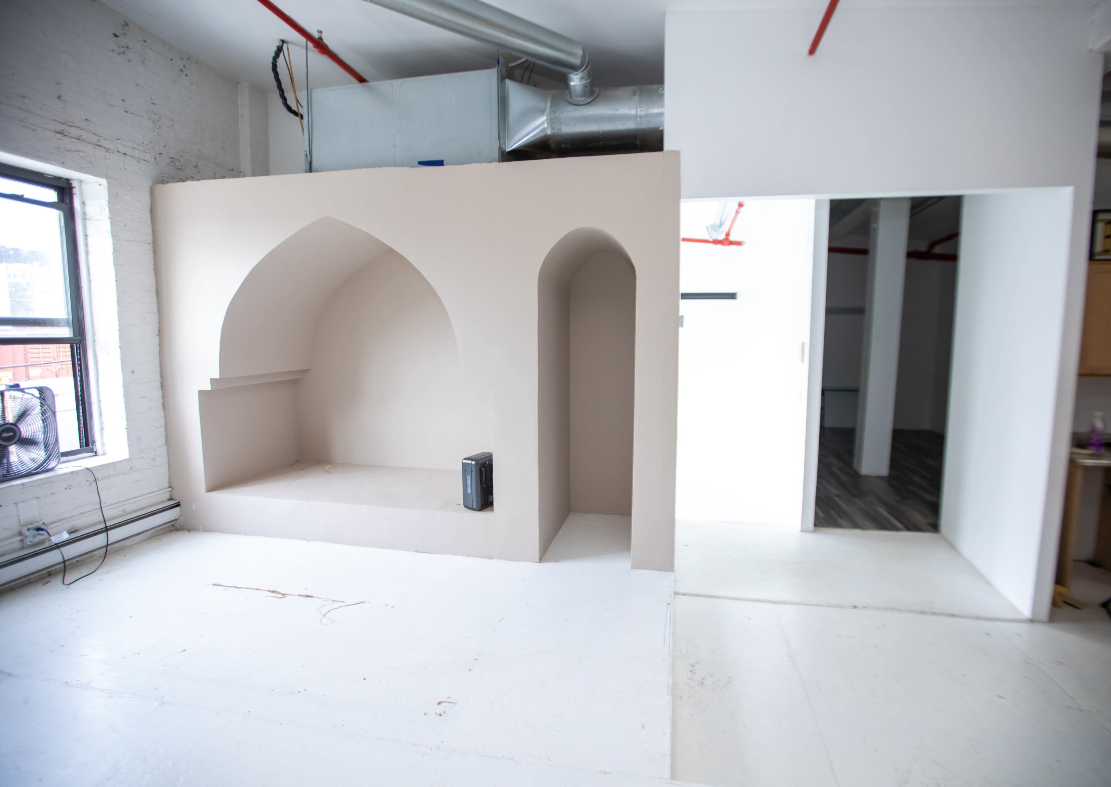 The image shows a minimalistic room featuring a curved alcove built into one wall, with a small black fan on a ledge and an open doorway leading to another space.