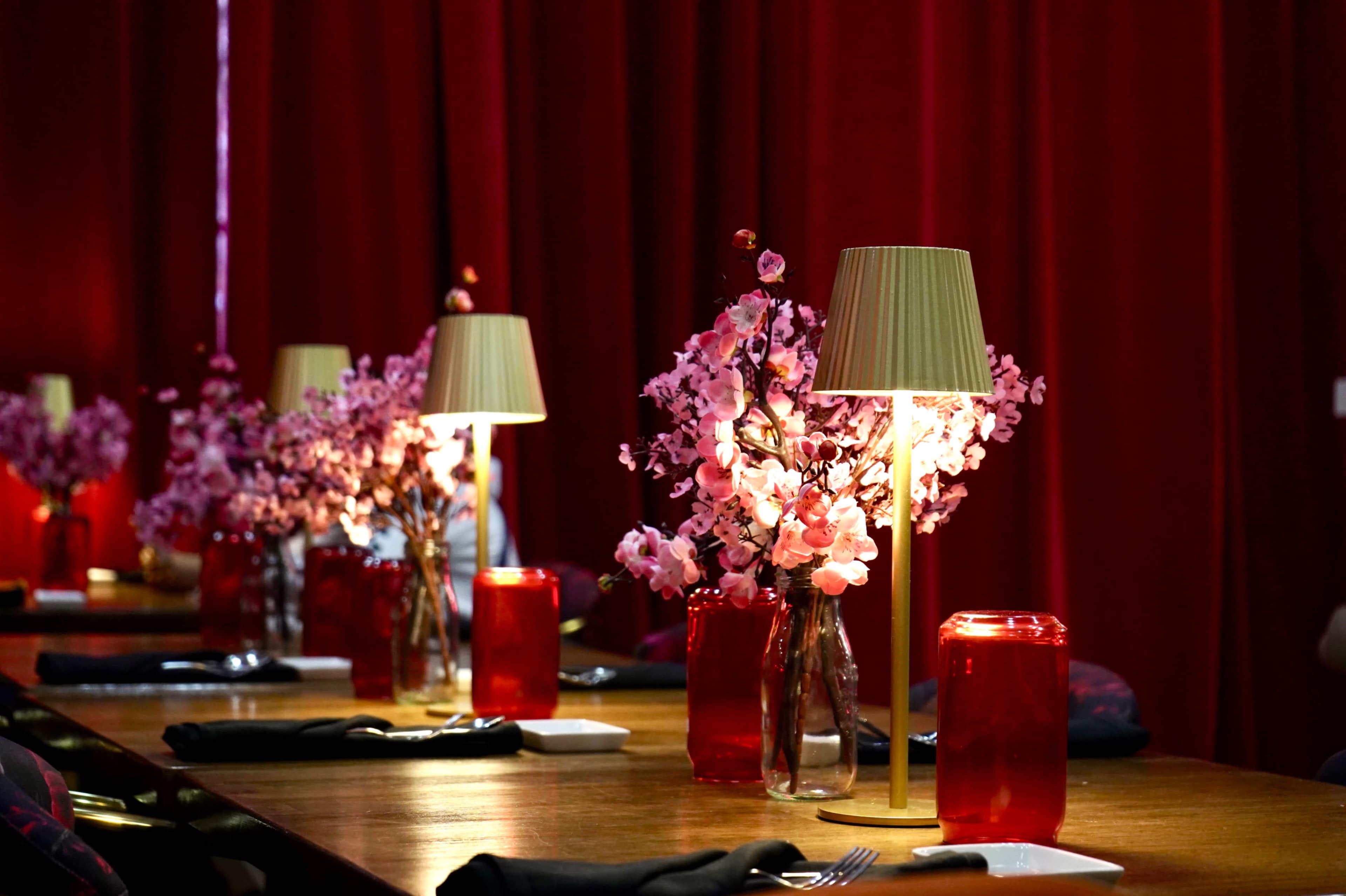 A long wooden table is set with red curtains in the background, adorned with pink flower arrangements and table lamps.