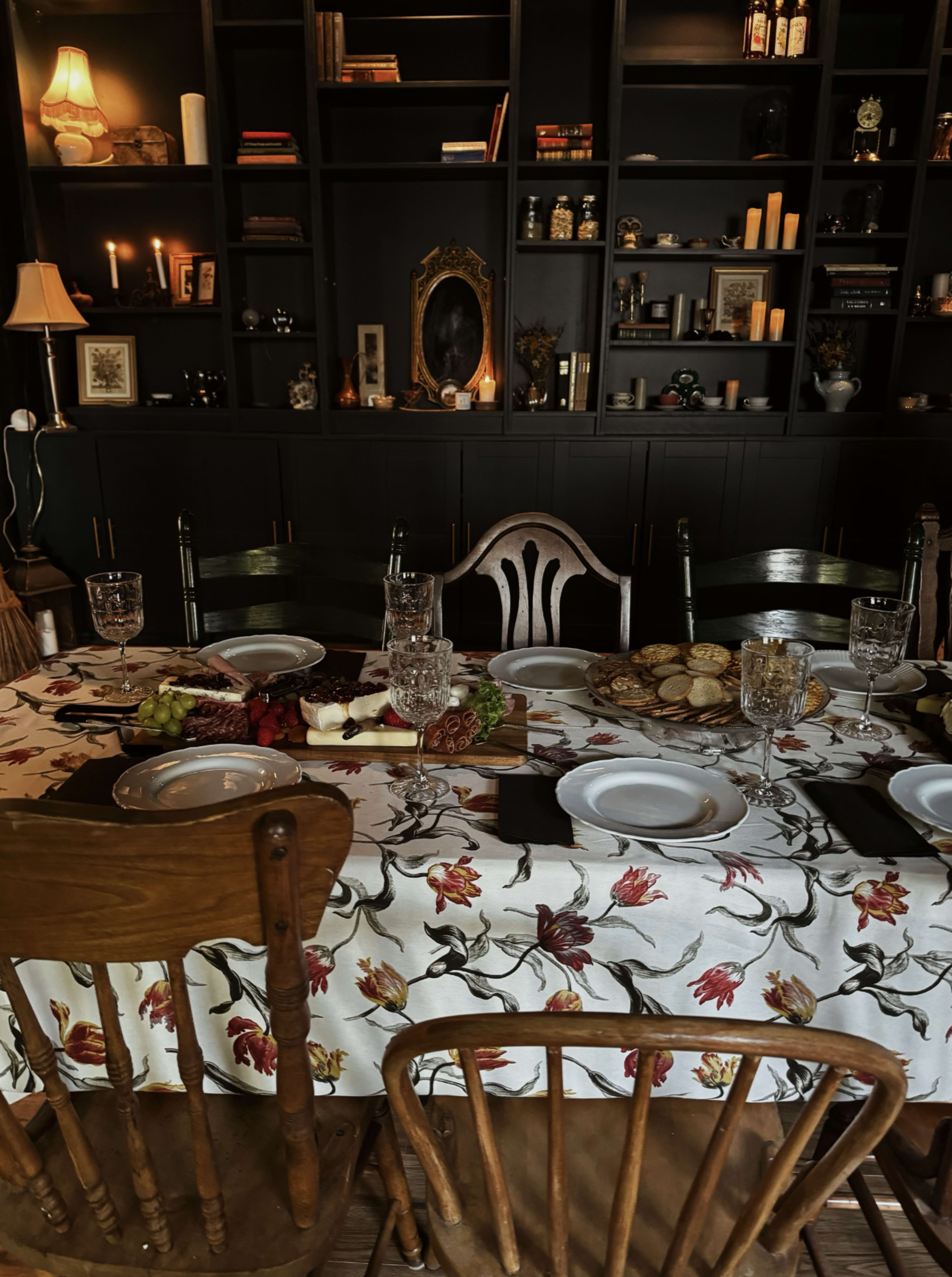 A dining table set with plates, glasses, and food is surrounded by wooden chairs in a room with dark shelves displaying various items.