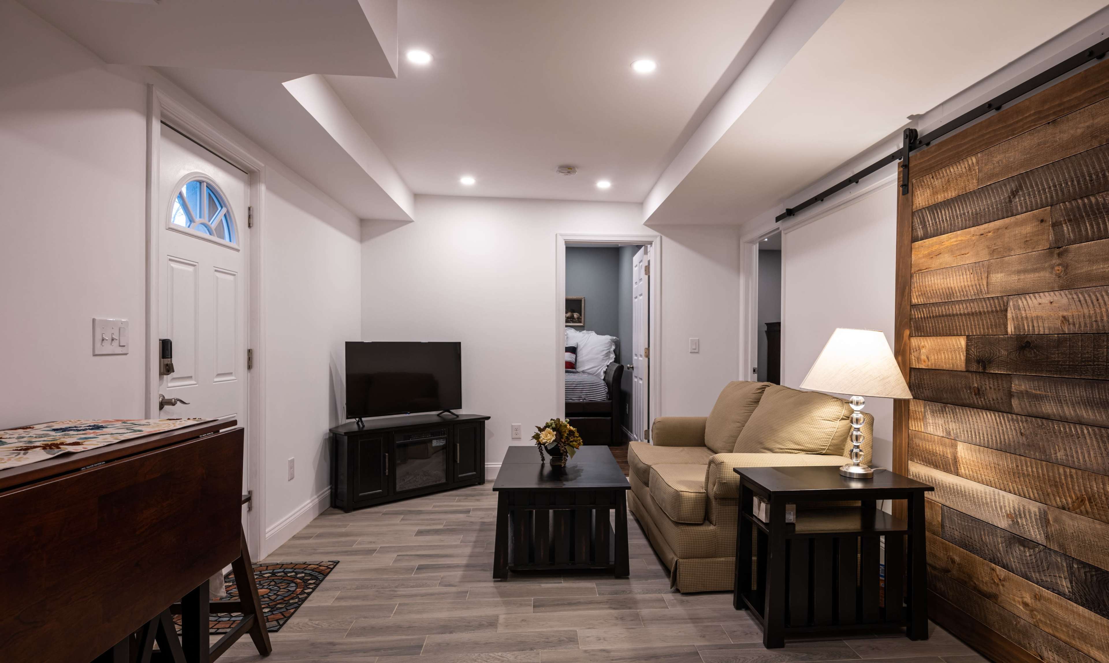 A small, modern living space features a beige couch, a television on a stand, and a wooden accent wall, with a doorway leading to a bedroom.