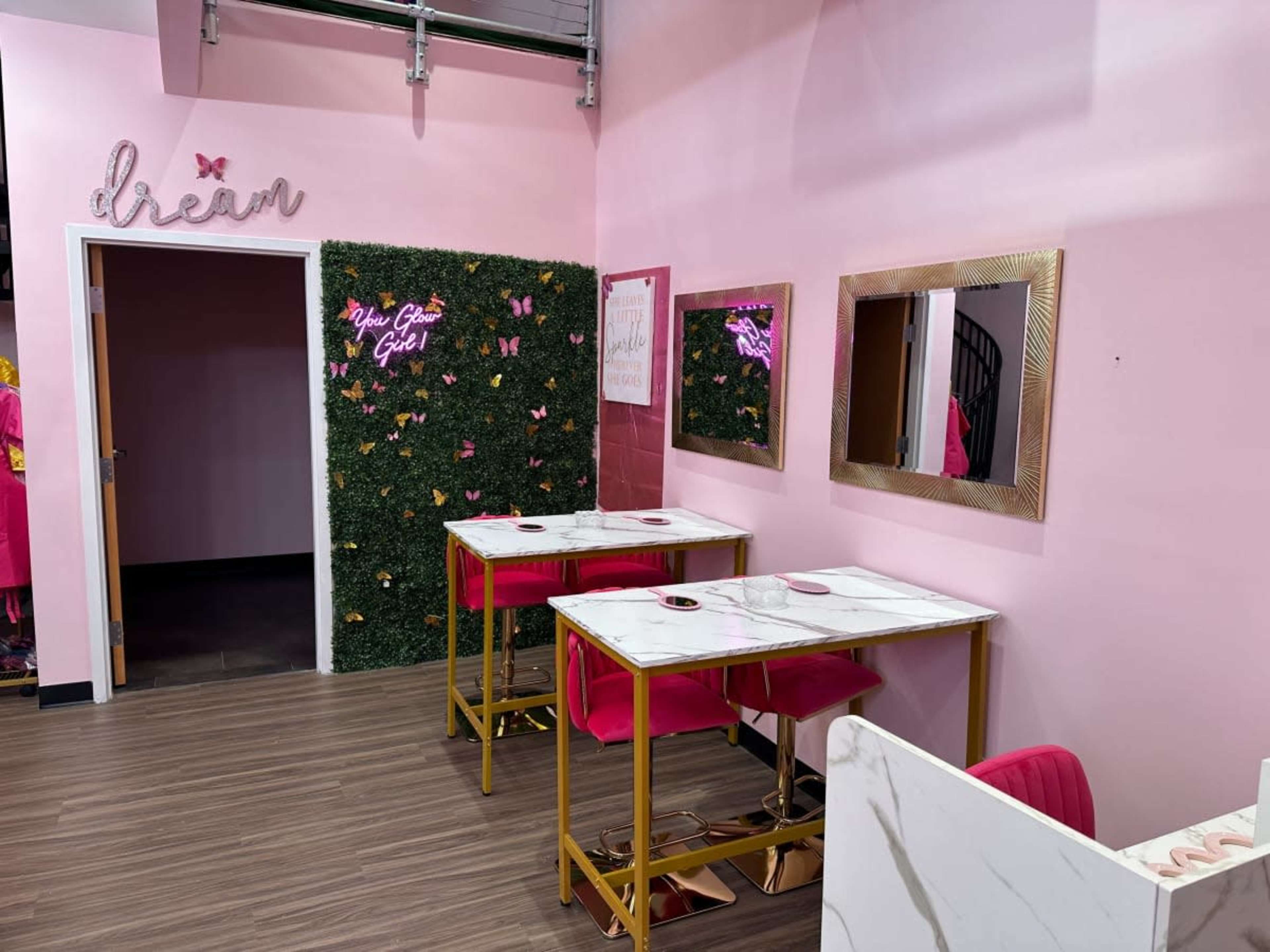 A brightly colored café interior featuring two marble-topped tables with pink chairs, a wall adorned with artificial greenery and butterflies, and framed mirrors.