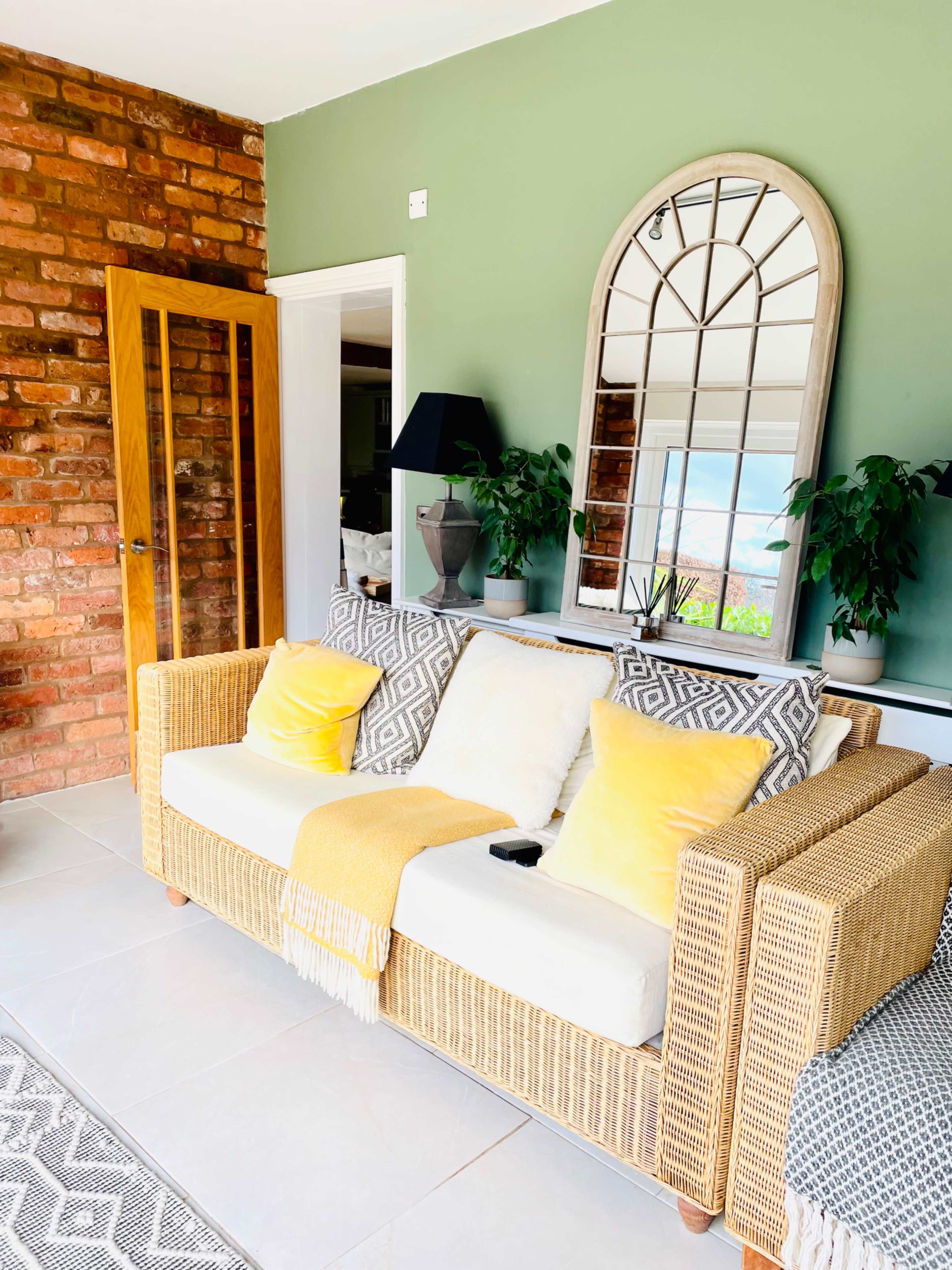 A wicker sofa with yellow pillows sits against a green wall, next to a mirror and plants in a well-lit room.
