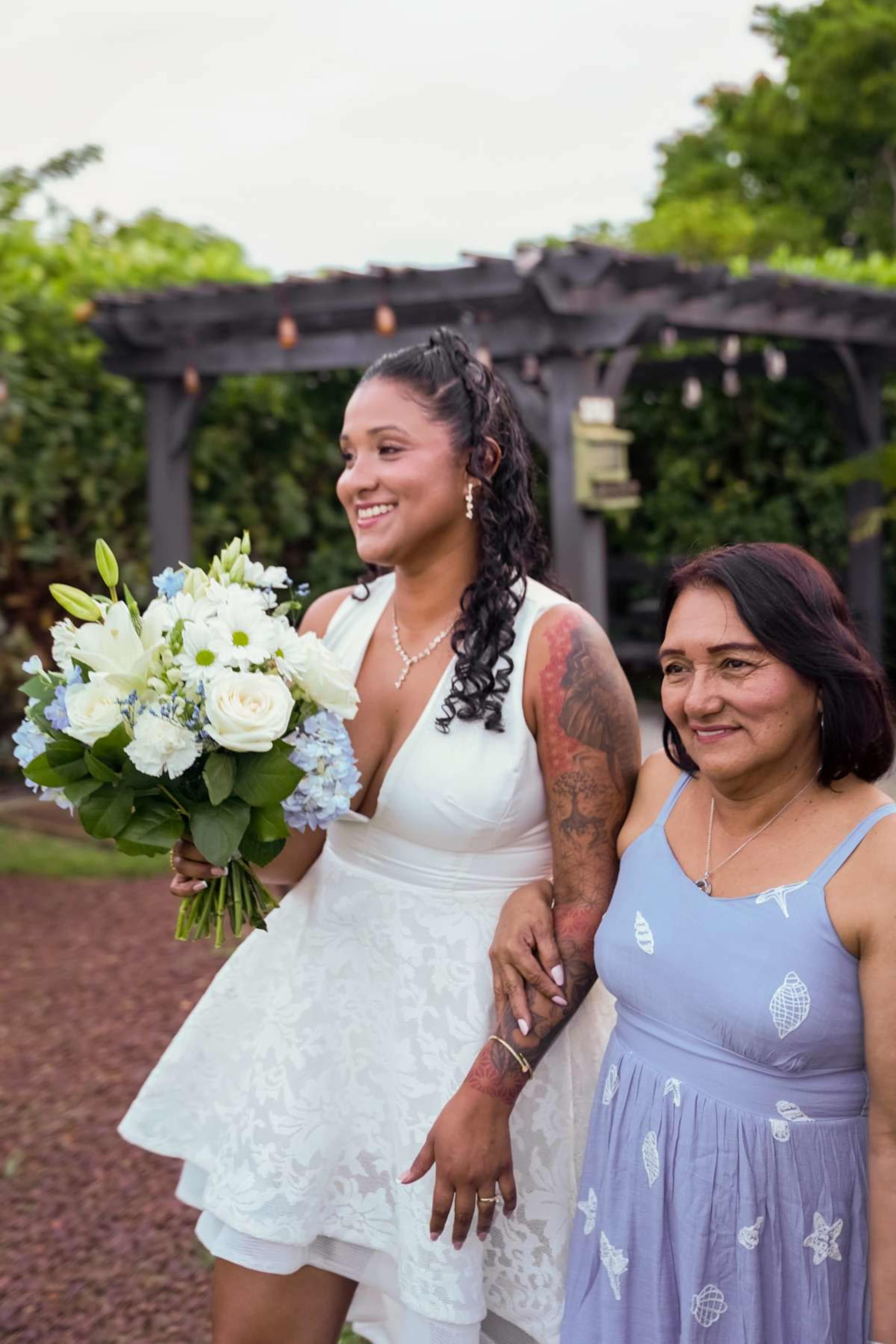 A bride in a white dress holds a bouquet of flowers while walking alongside a woman in a light blue dress.