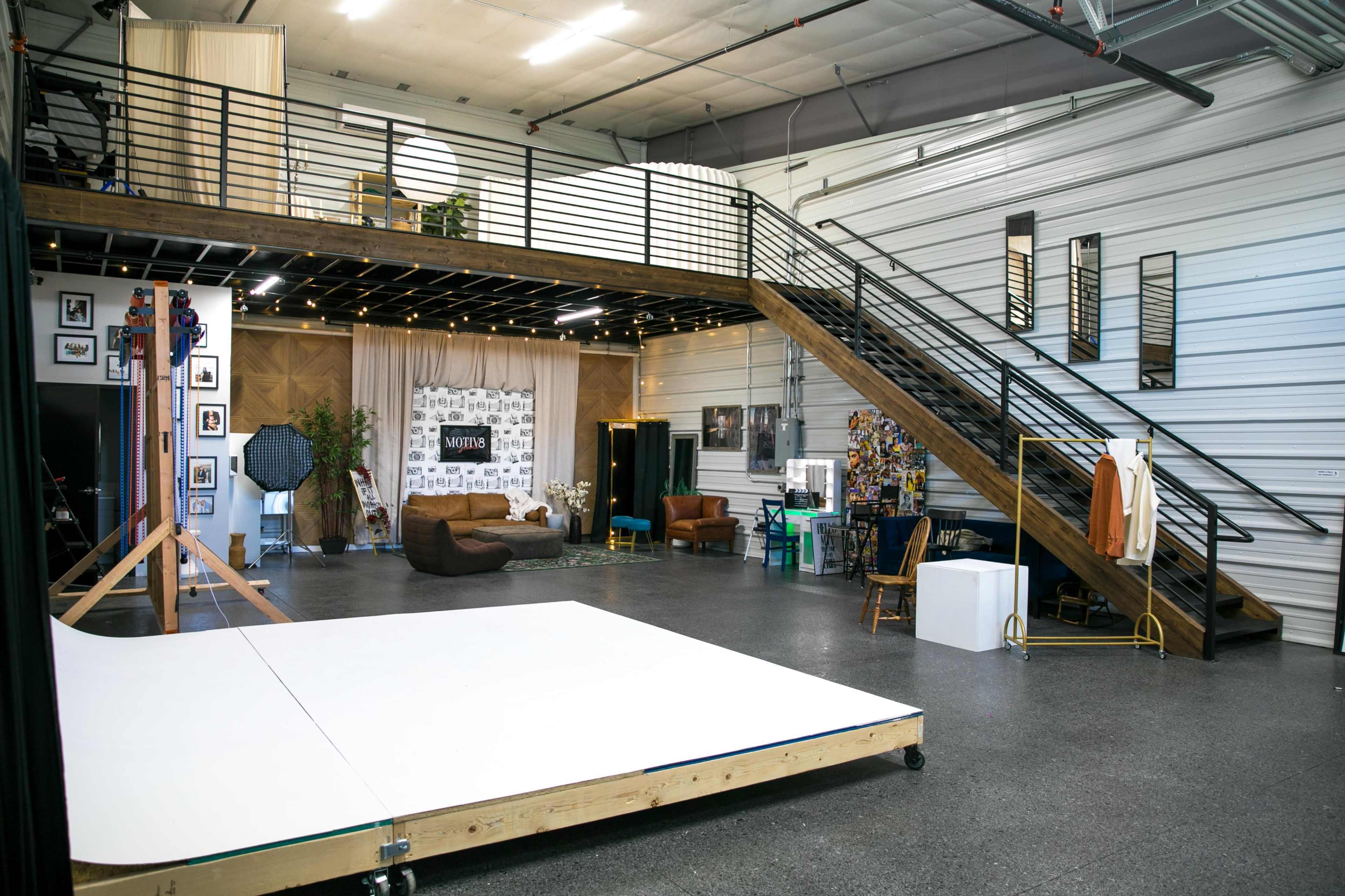 The image shows a spacious studio featuring a wooden lofted area, a white platform, various seating arrangements, and decorative elements against a backdrop of metallic walls.