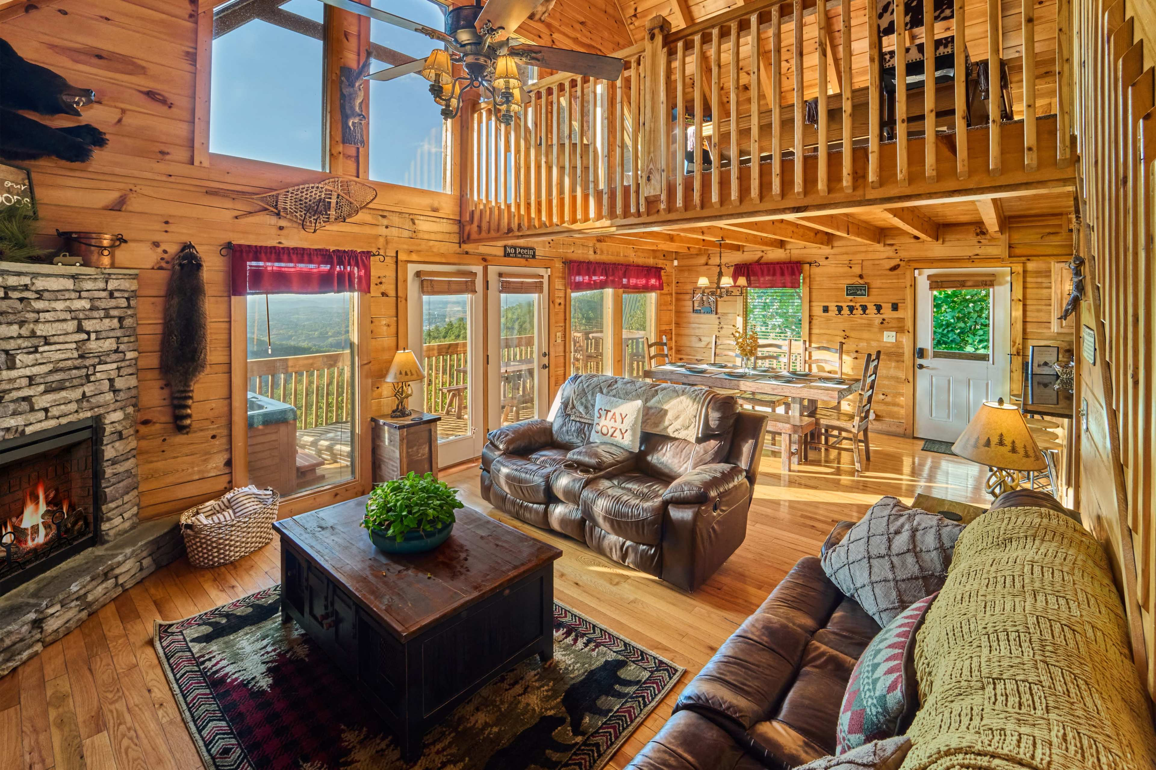 The image shows a spacious wooden cabin interior with a fireplace, leather seating, a dining area, and large windows that allow natural light to fill the room.