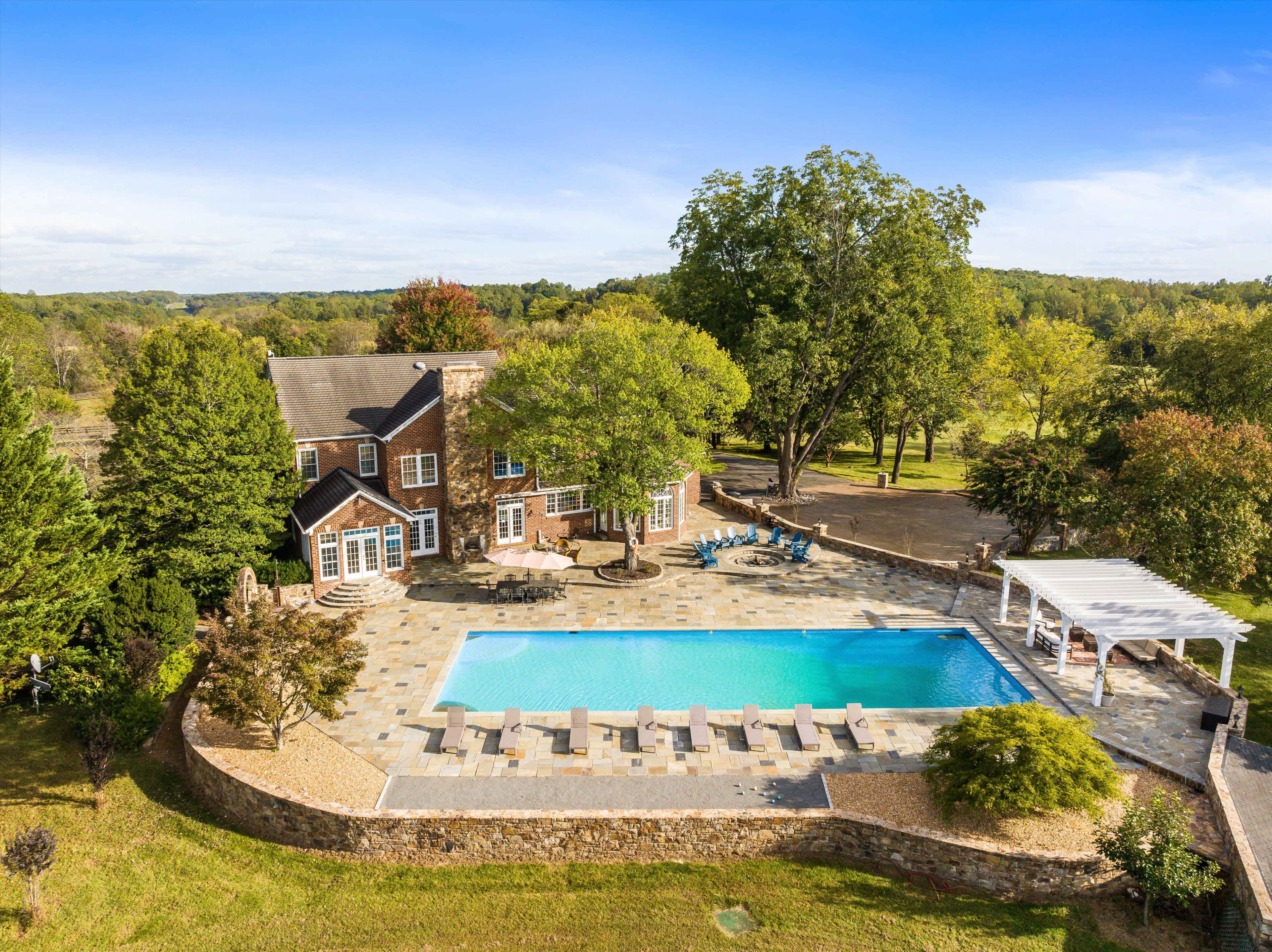 The image shows a large house adjacent to a swimming pool surrounded by landscaped grounds and trees.