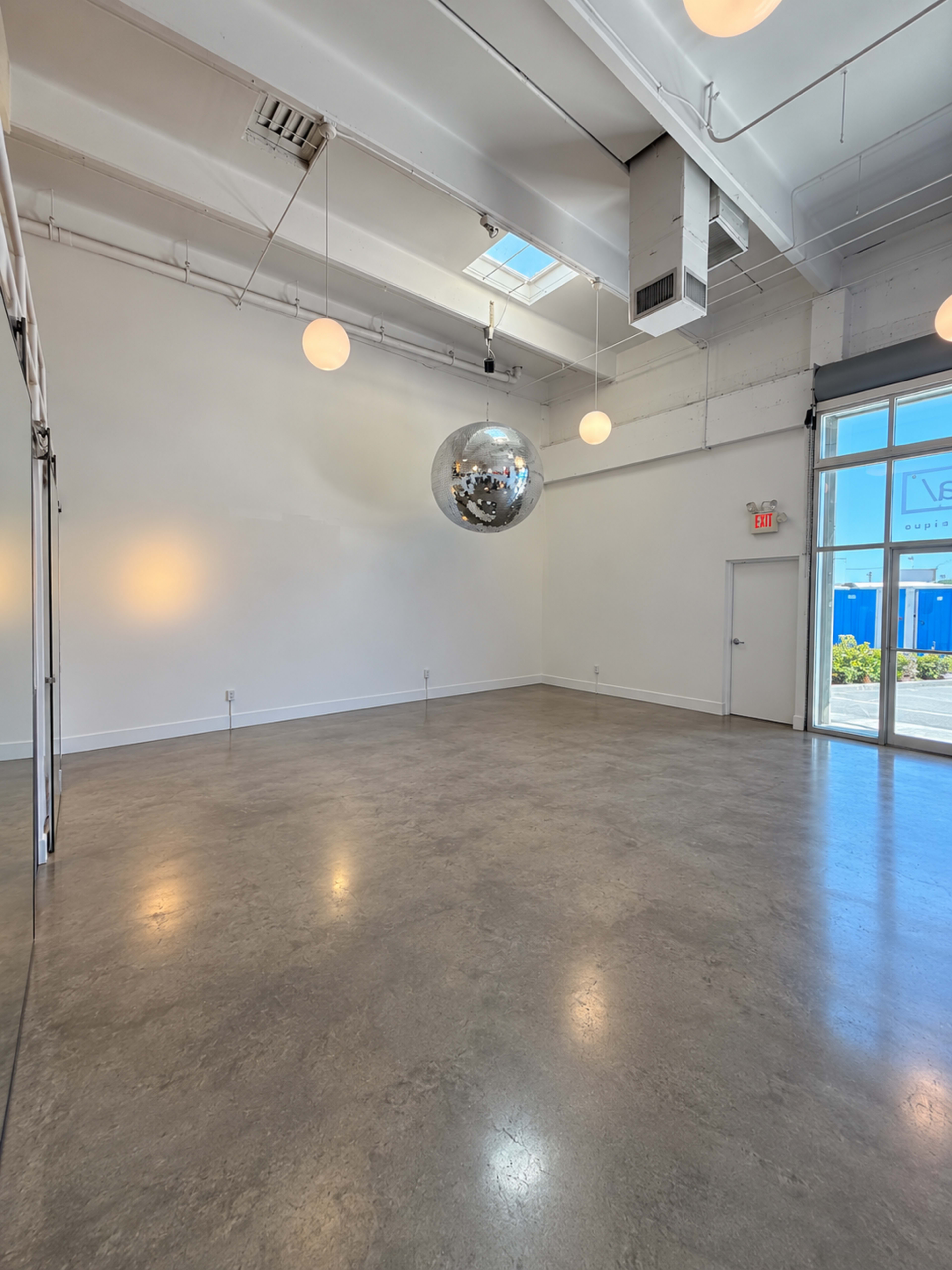 The image shows a spacious, empty room with a polished concrete floor, featuring a large disco ball suspended from the ceiling and a glass entrance leading outside.