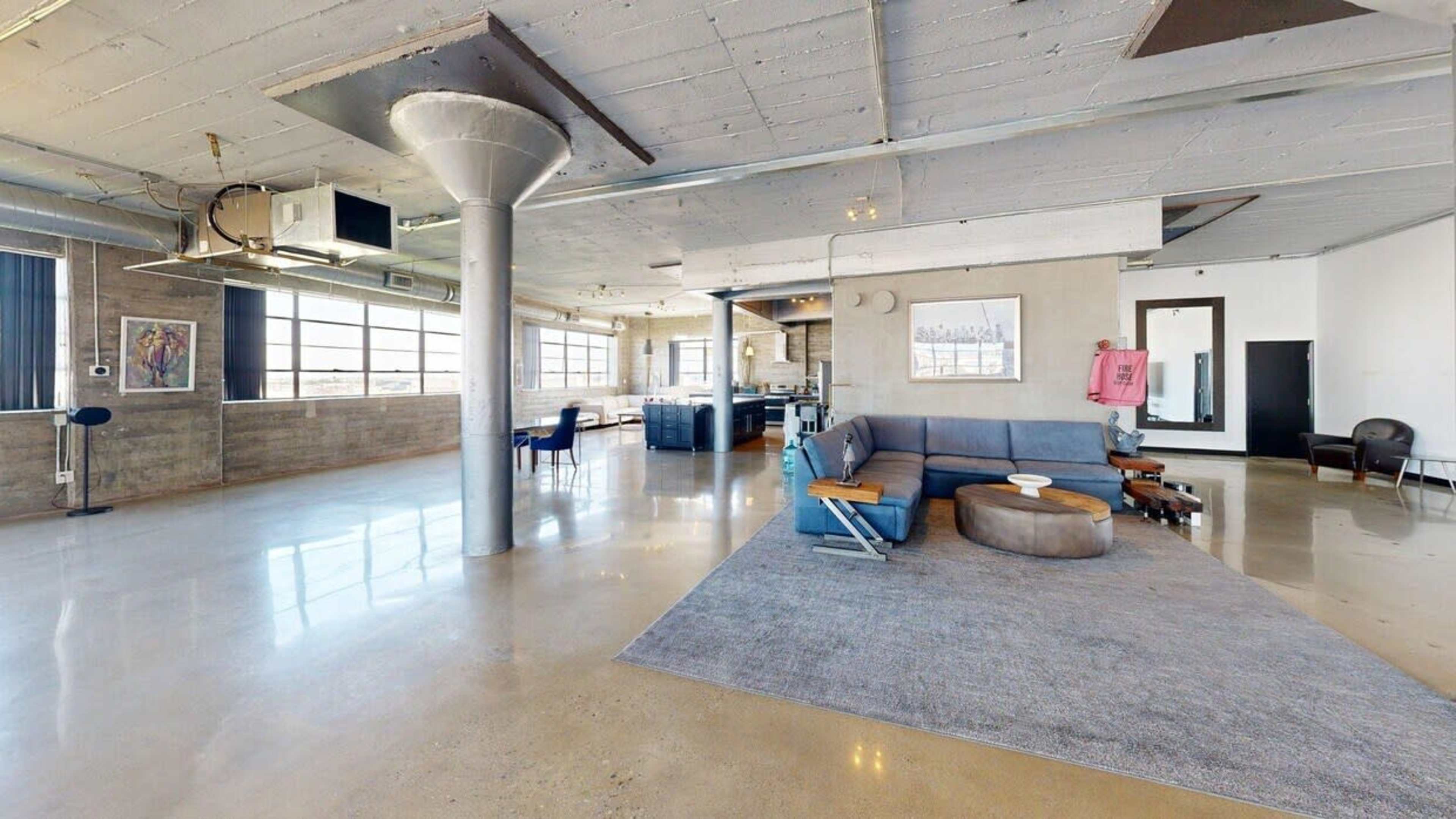 The image shows a spacious, modern interior with large windows, polished concrete flooring, a sectional sofa, and various seating areas.