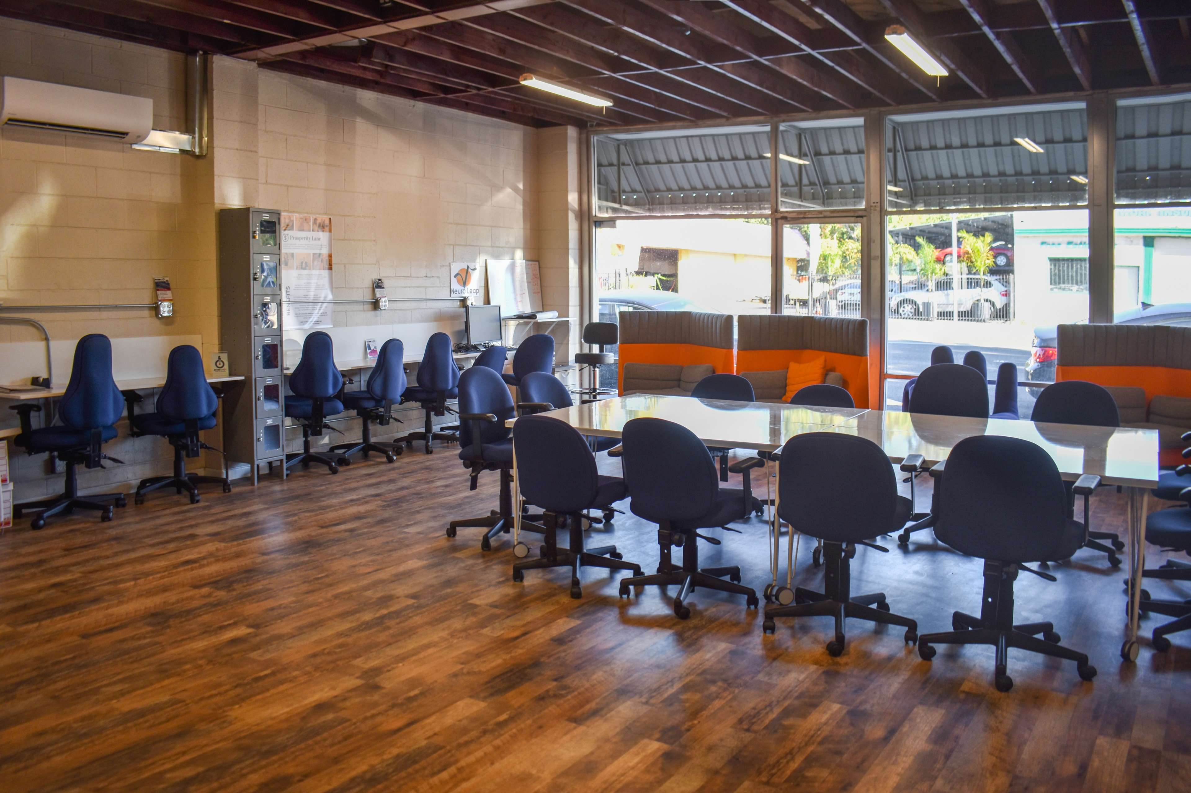 The image shows a meeting room with several blue chairs arranged around a large table and large windows letting in natural light.