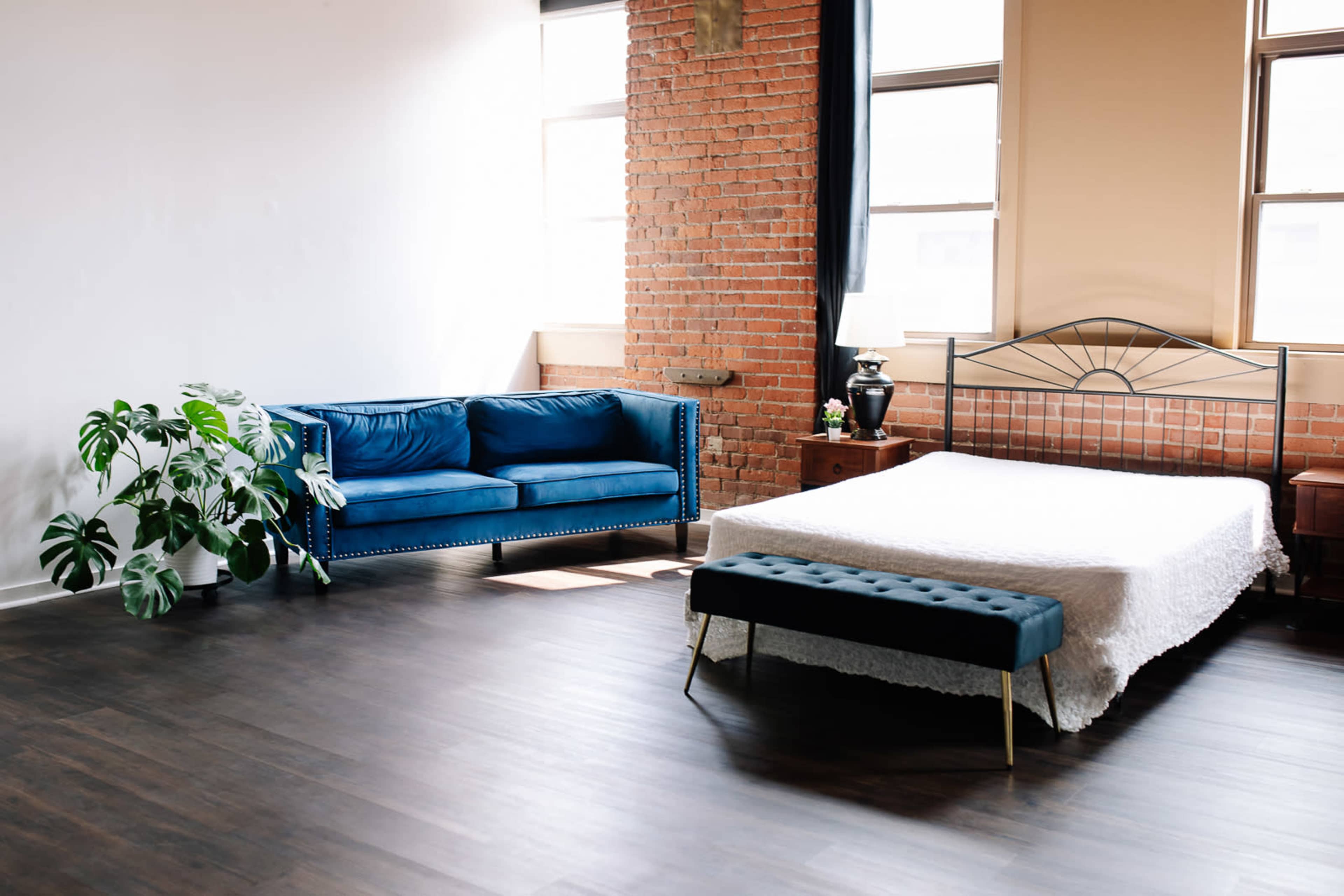 A spacious bedroom features a blue couch, a white bedspread over a bed, and a plant in a well-lit room with exposed brick walls.