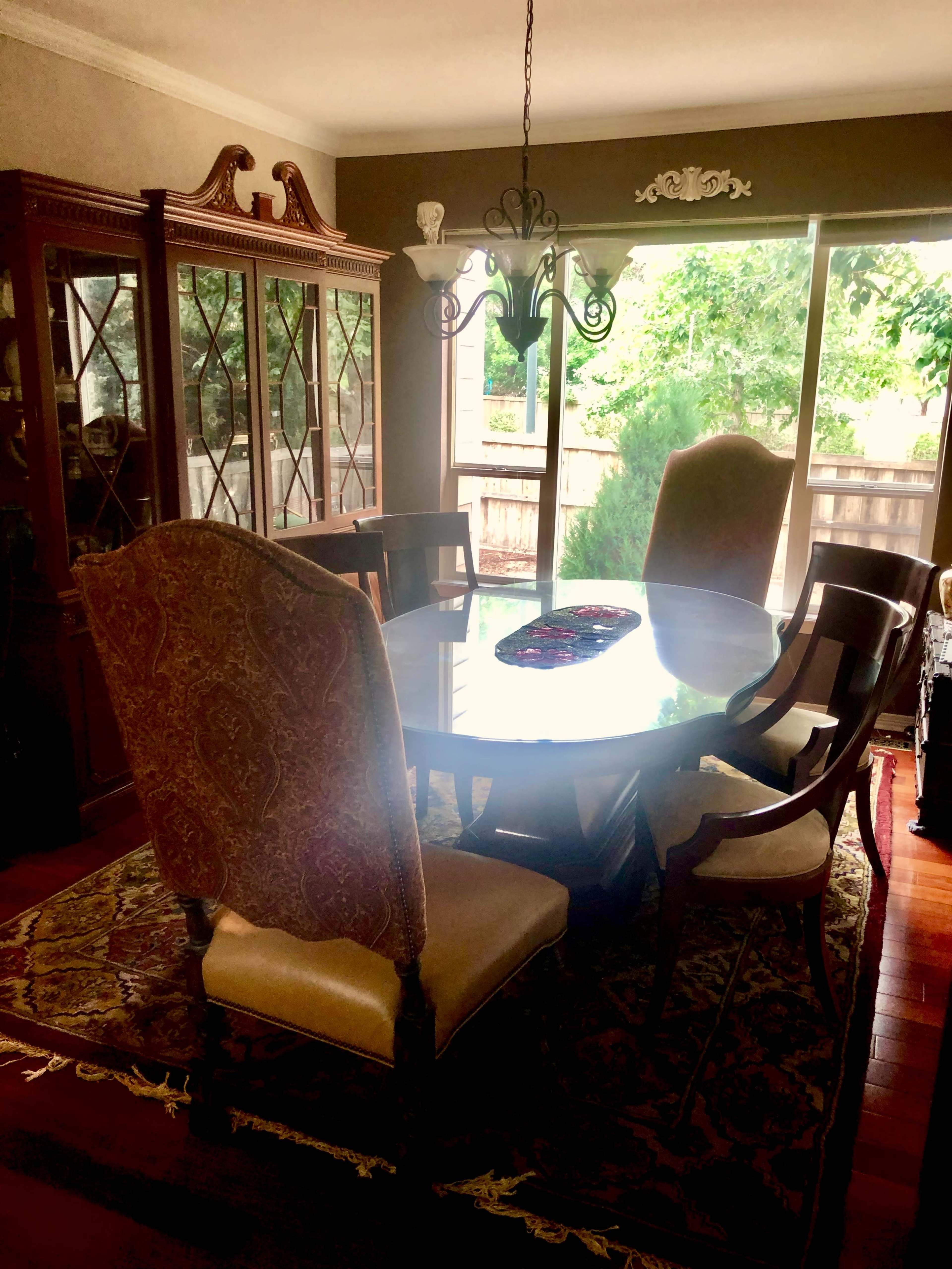 A dining room with a round table surrounded by six chairs, a china cabinet in the corner, and a window letting in natural light.
