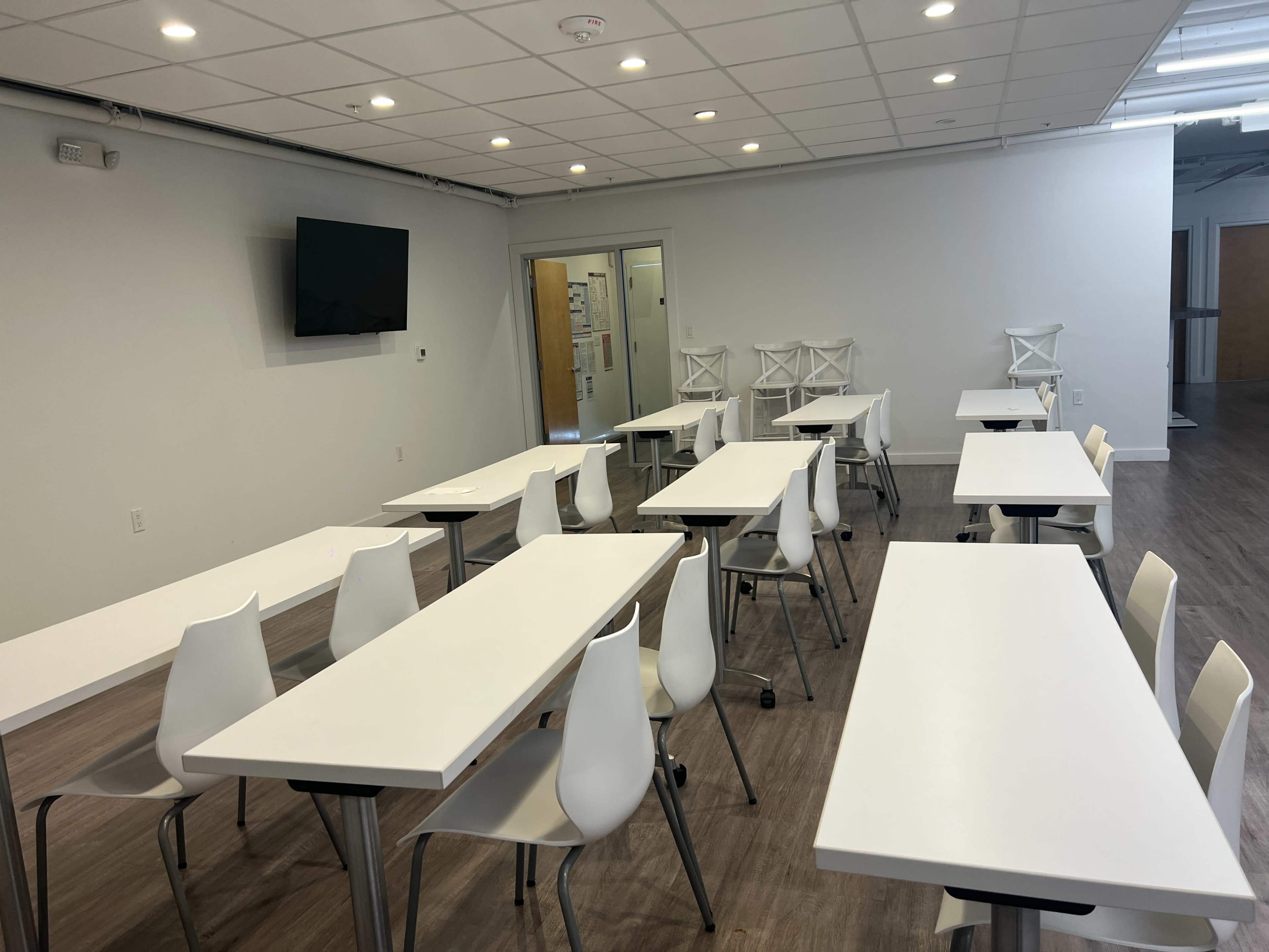 A modern classroom with multiple white tables and chairs arranged to face a wall-mounted television.