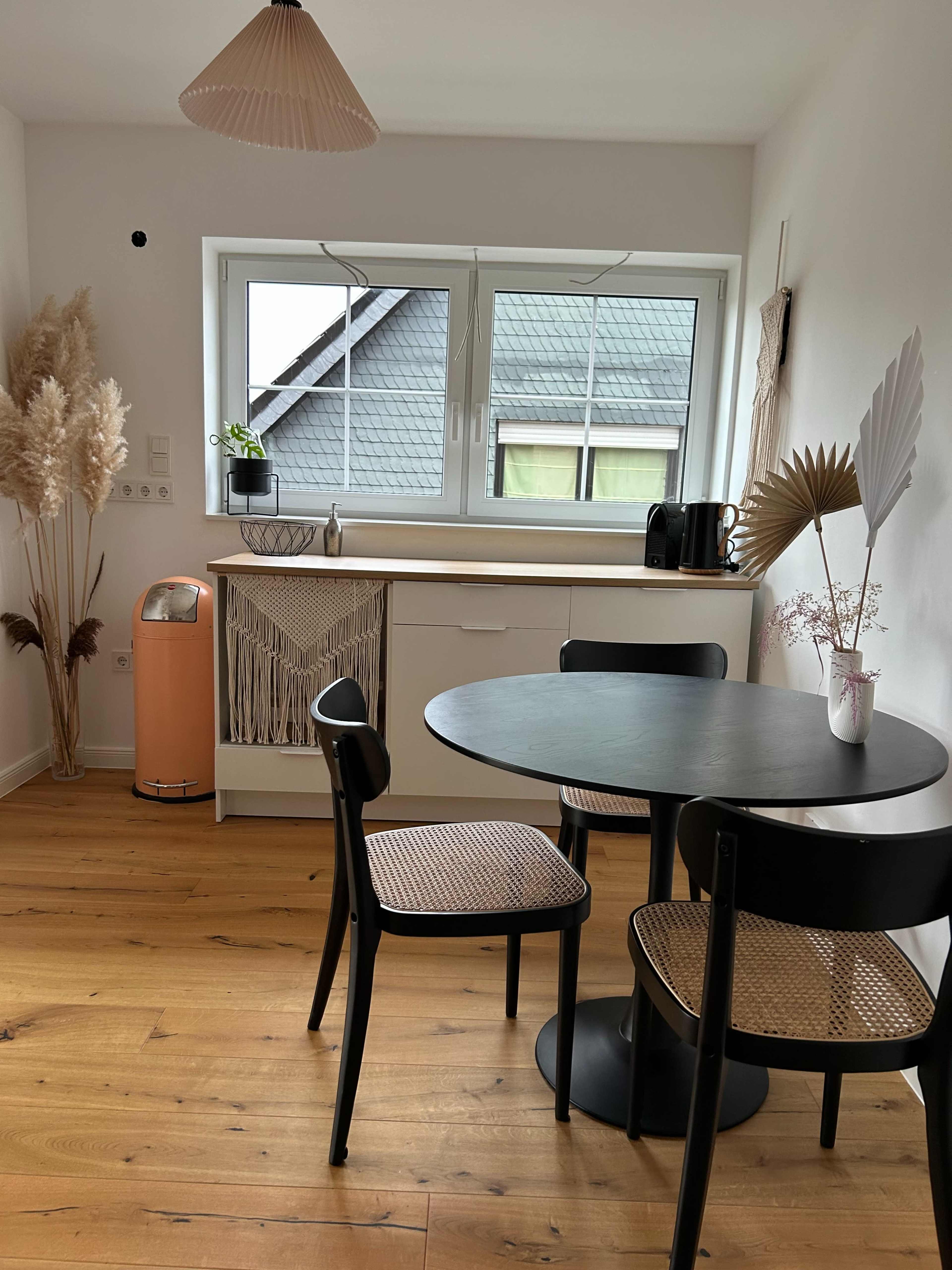 A modern kitchen features a round black table with two chairs, a light-colored cabinet, an orange kitchen appliance, and decorative plants by the window.