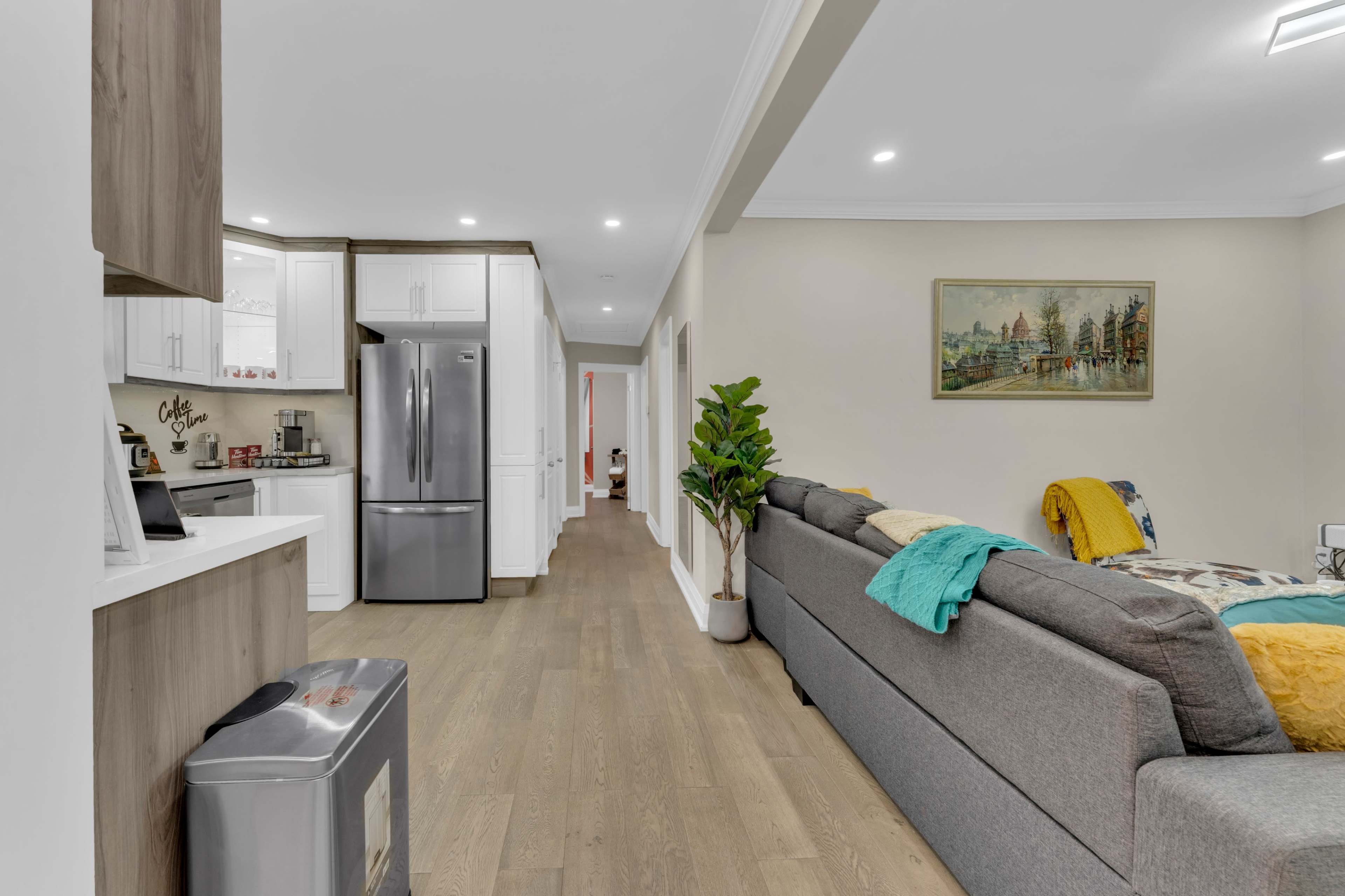 A modern kitchen and living room area with a gray sofa, a refrigerator, and a long hallway leading to additional rooms.