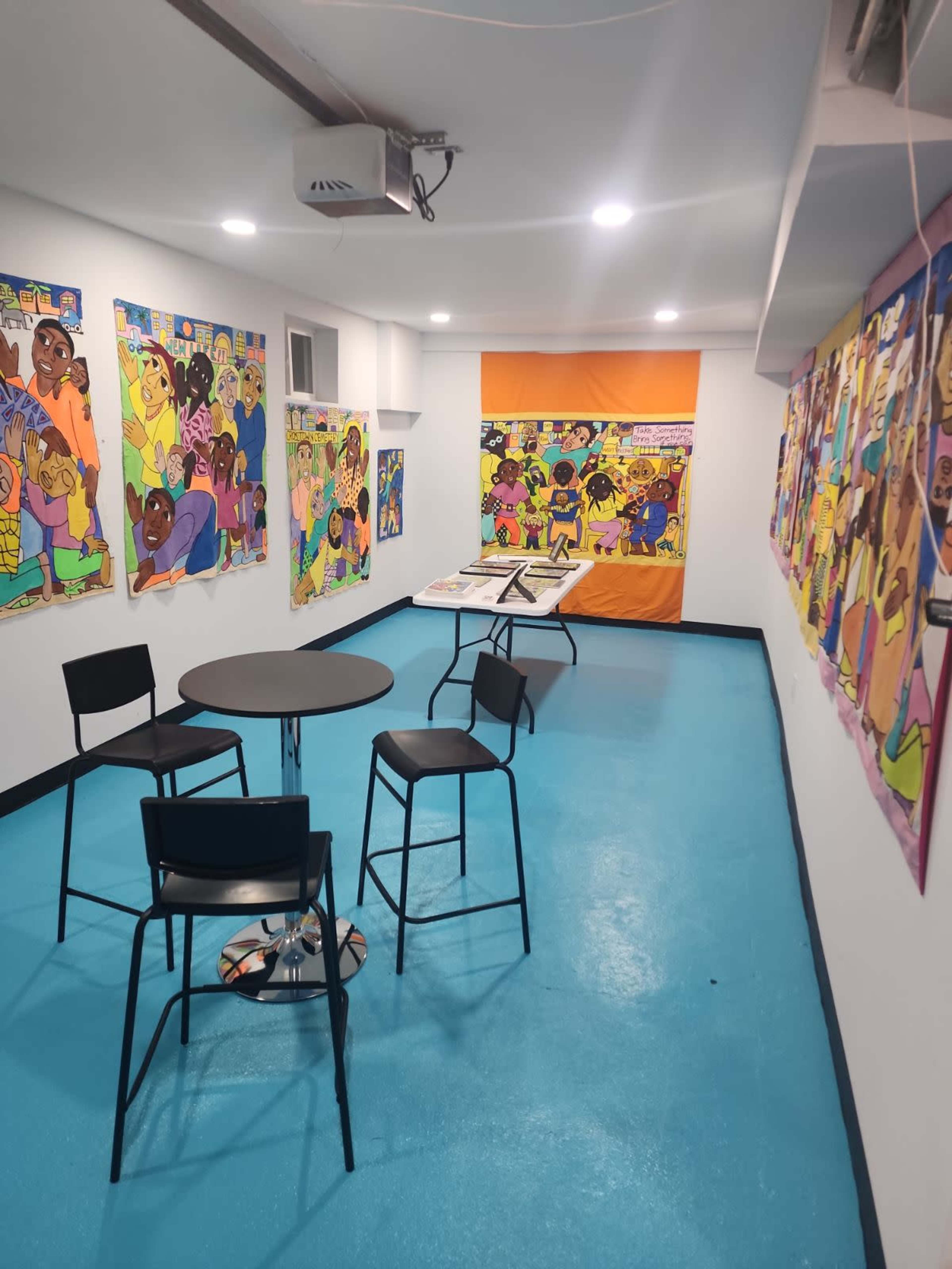 The image shows a brightly lit room with colorful wall art, a round table, and four black chairs arranged on a blue floor.