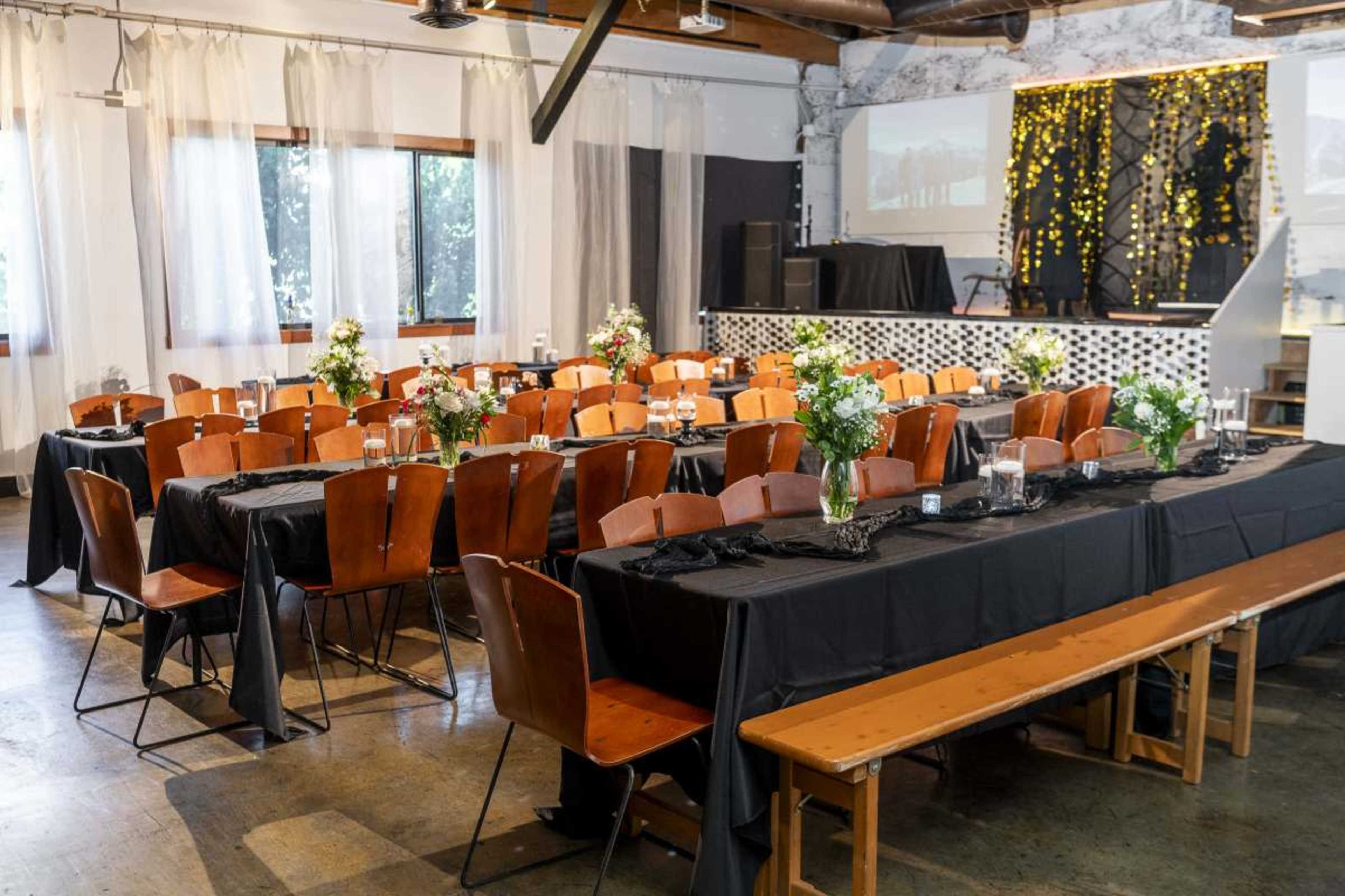 The image shows an event space arranged with multiple tables set with black tablecloths and floral centerpieces, ready for a gathering.