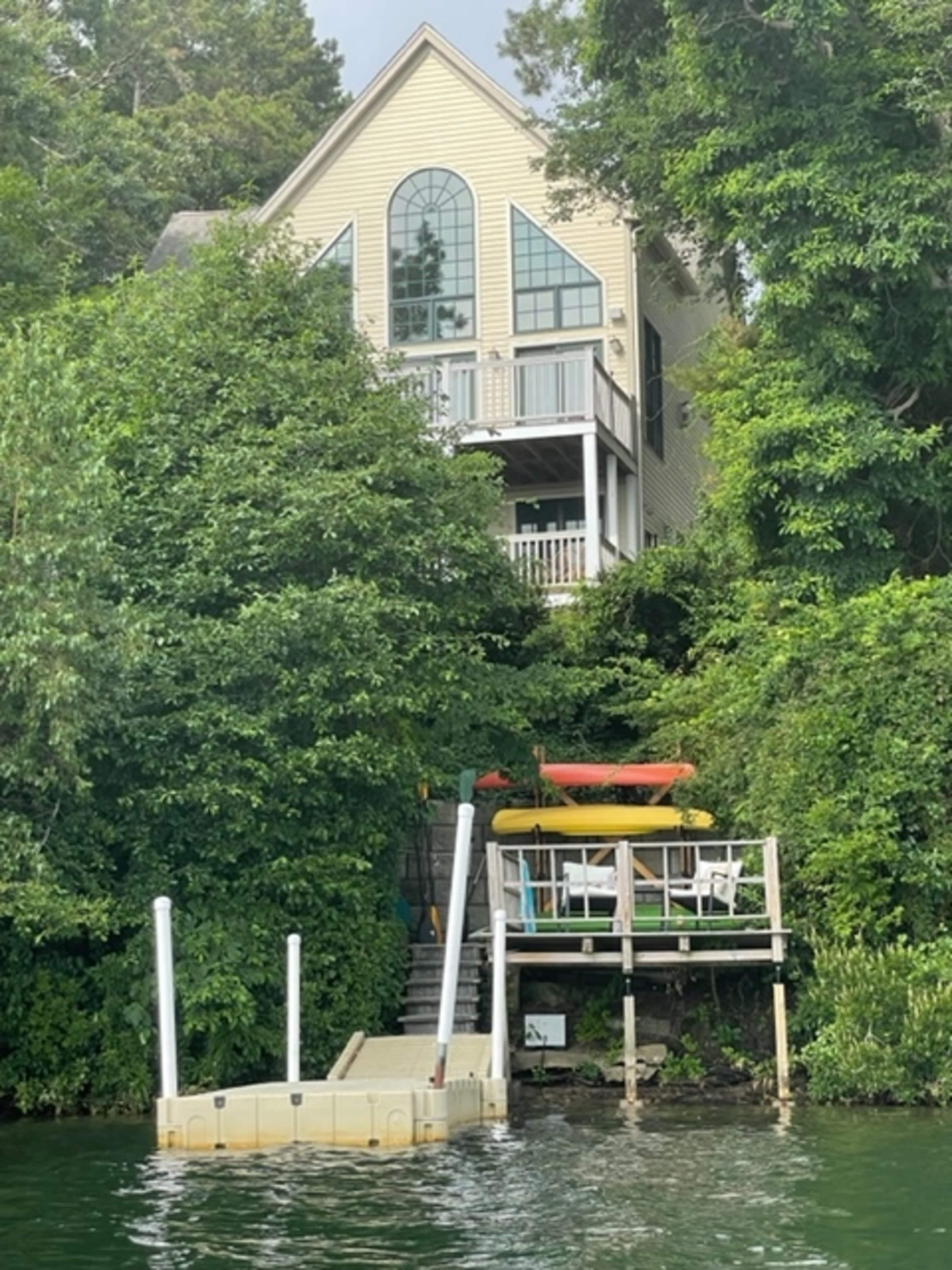 A two-story yellow house with large windows sits on a hillside, overlooking a dock and kayaks by the water, surrounded by lush greenery.
