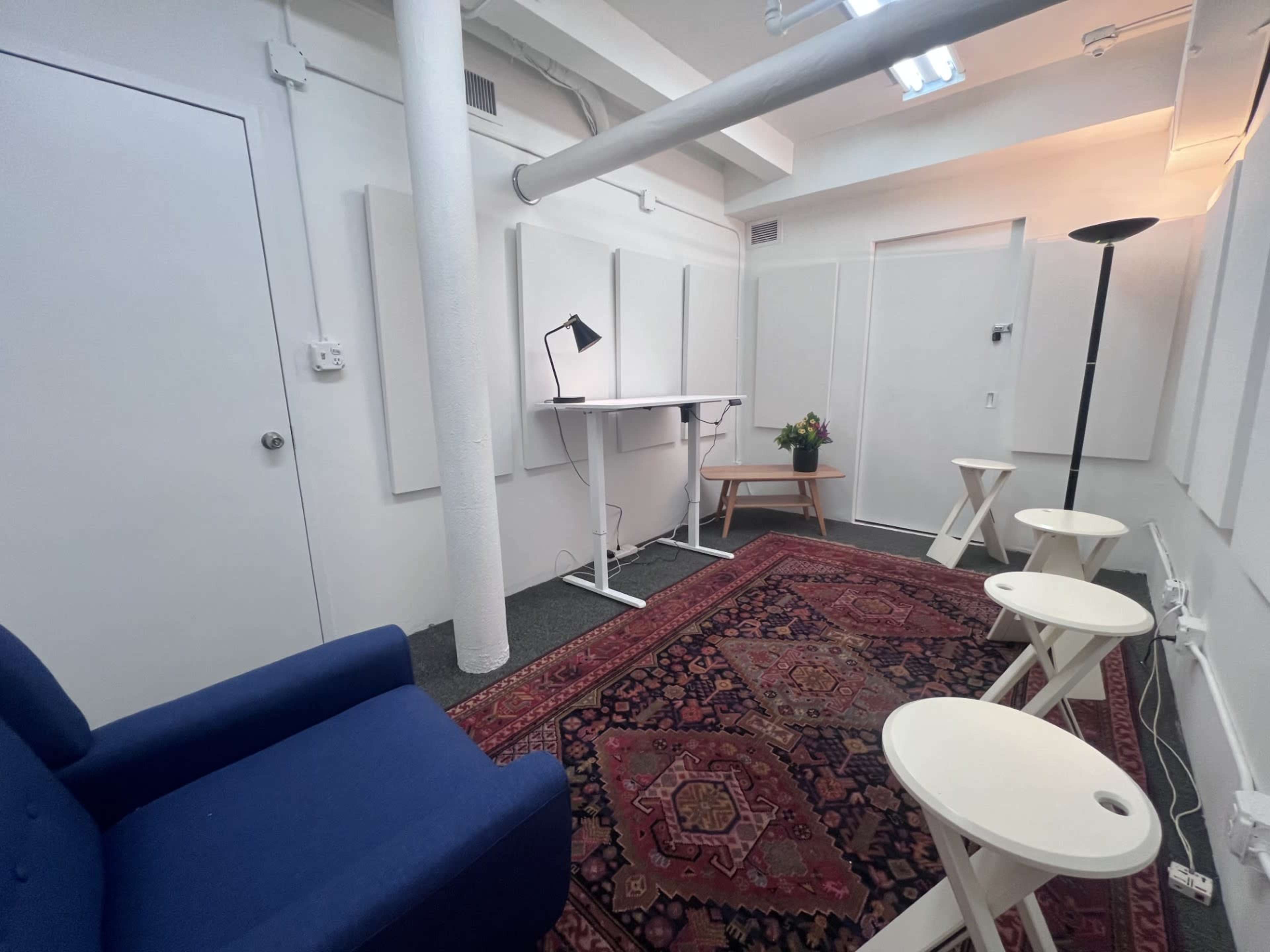 The image shows a small, modern workspace featuring a blue chair, a white standing desk, several small white side tables, and a floral arrangement on a wooden table, all set against a patterned rug.