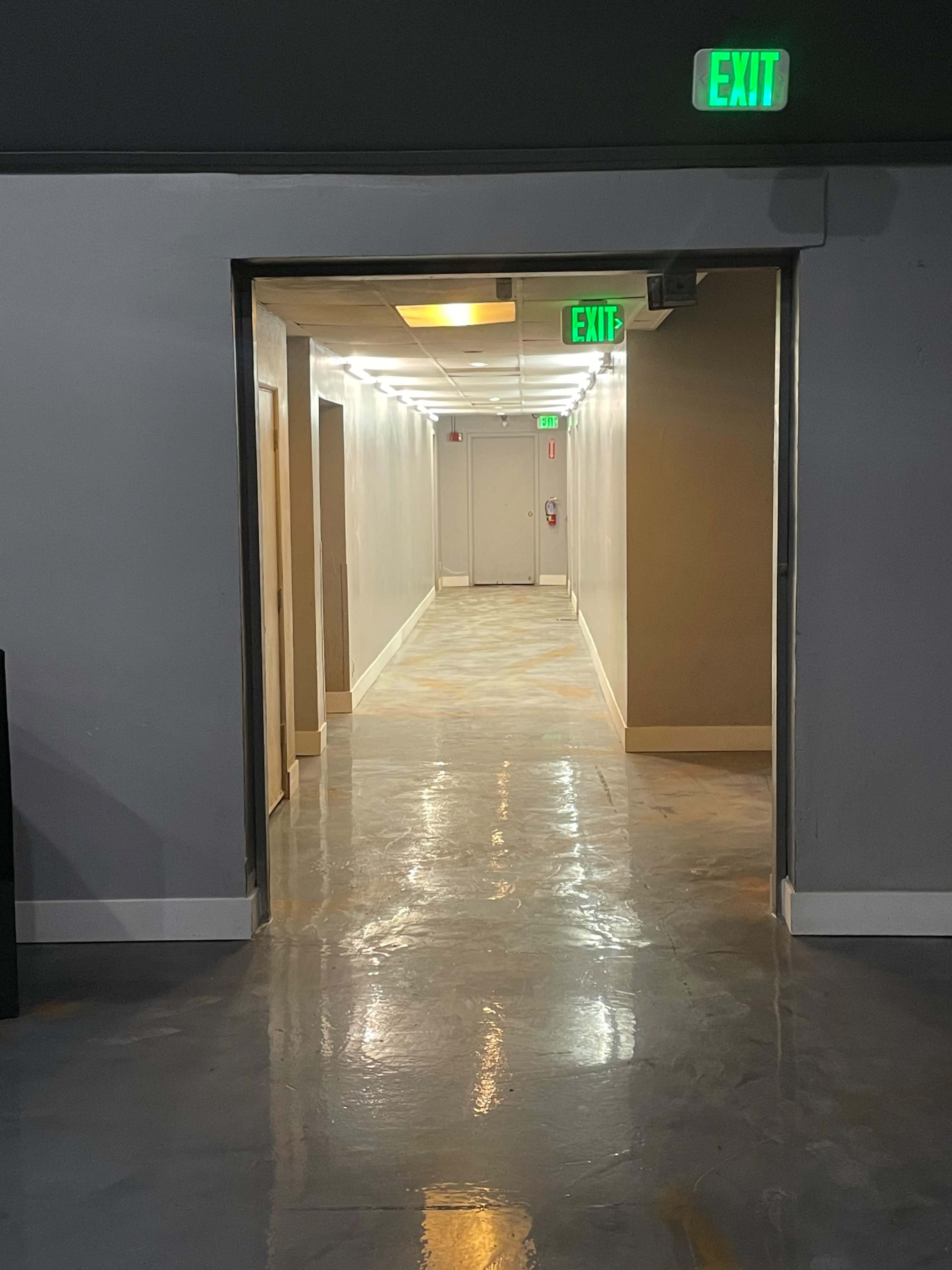 A long hallway with gray walls and a shiny floor leads to an exit door at the end, illuminated by green exit signs.