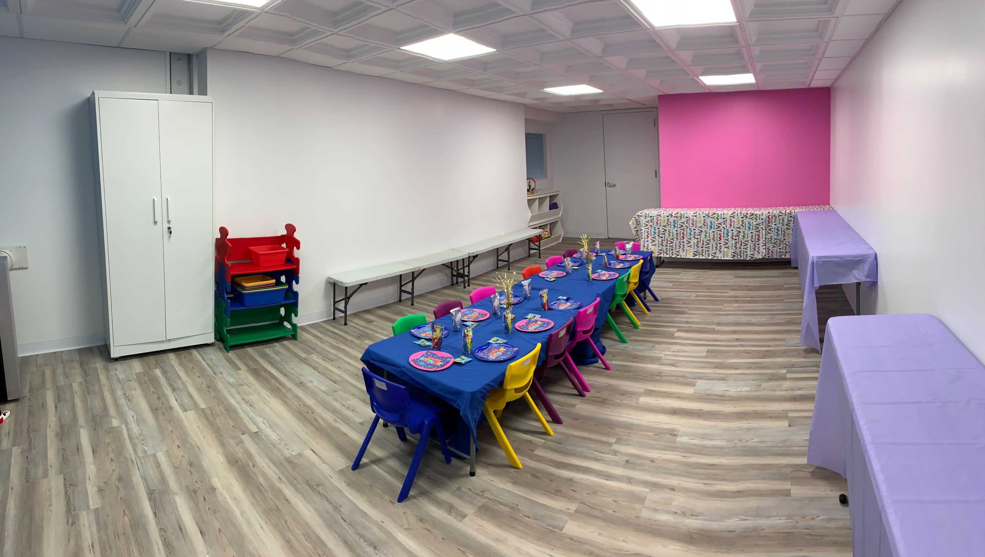 The image shows a brightly colored children's party room featuring a long table set for a celebration with colorful chairs, along with a storage cabinet and a decorated table in the background.