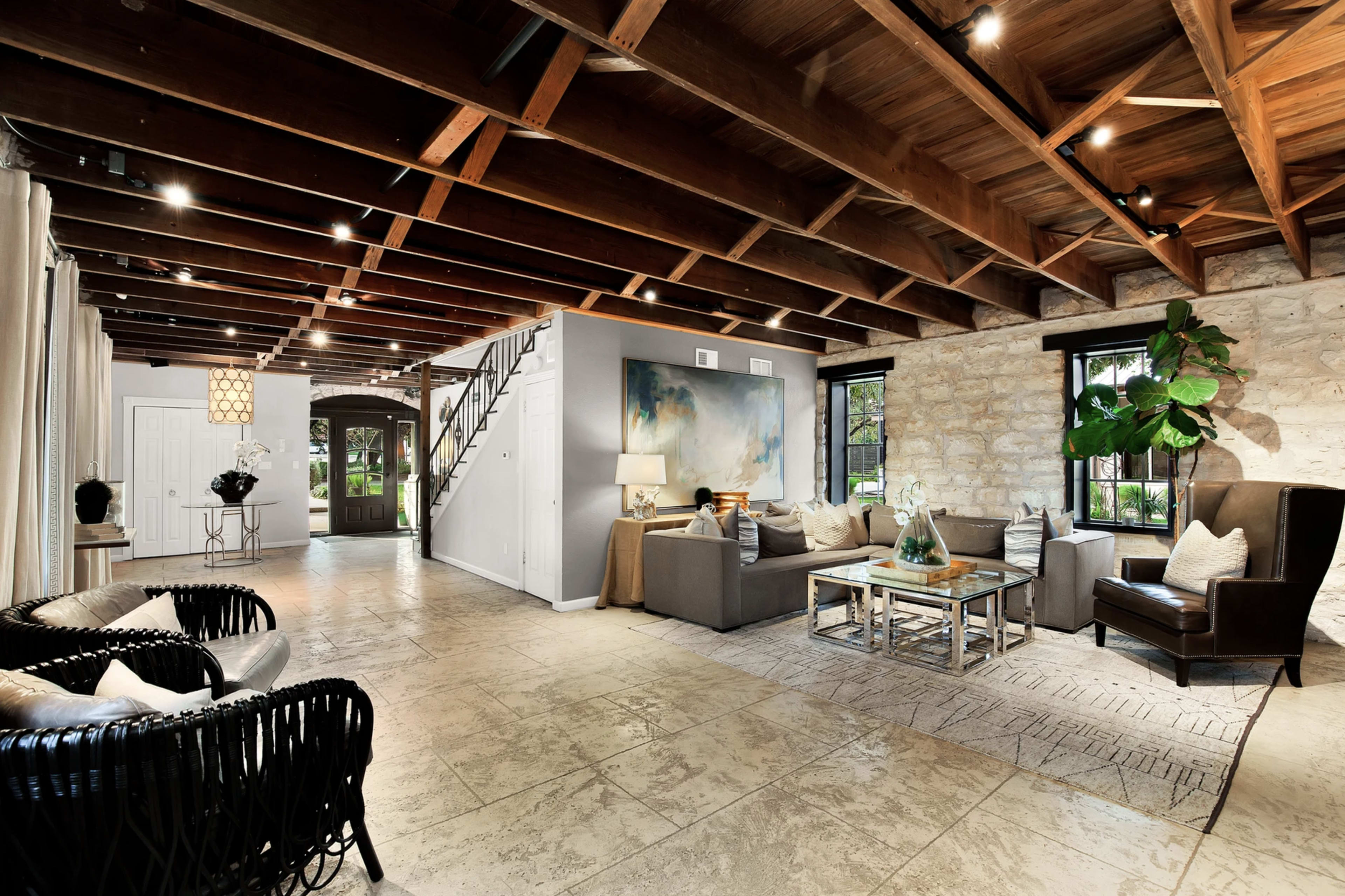 The image shows a spacious living room with a high wooden ceiling, stone walls, and a combination of modern and traditional furniture, including a sofa set, an armchair, and a coffee table.