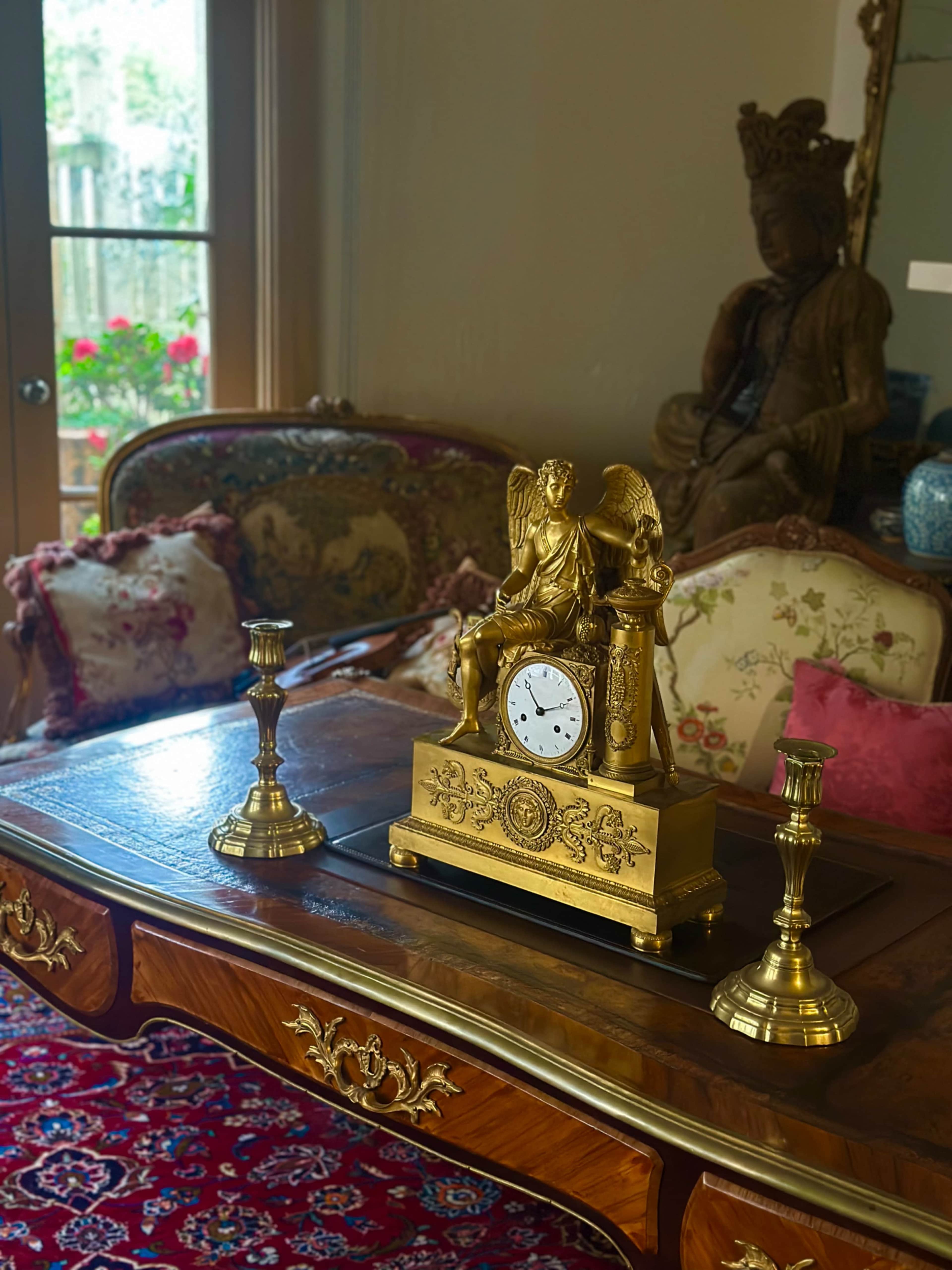 A vintage golden clock featuring a winged figure is placed on a wooden table between two candlesticks in a decorated room.