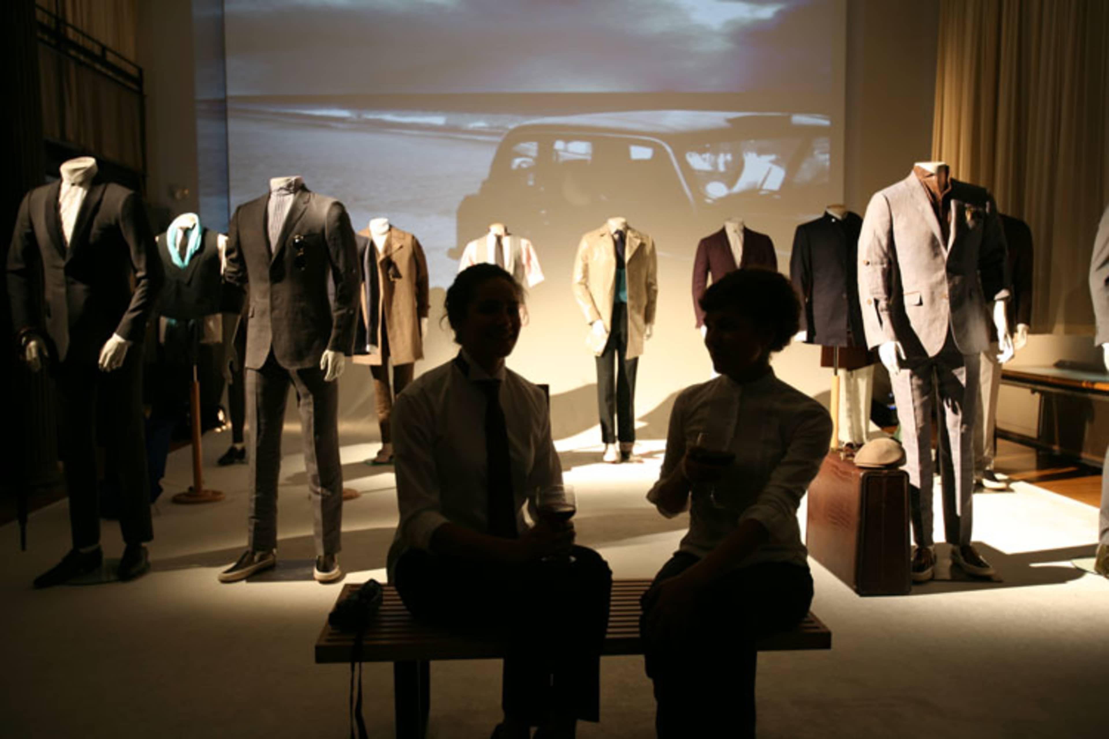 Two individuals sit on a bench in front of a display of various suits and an image of a car projected on a wall.
