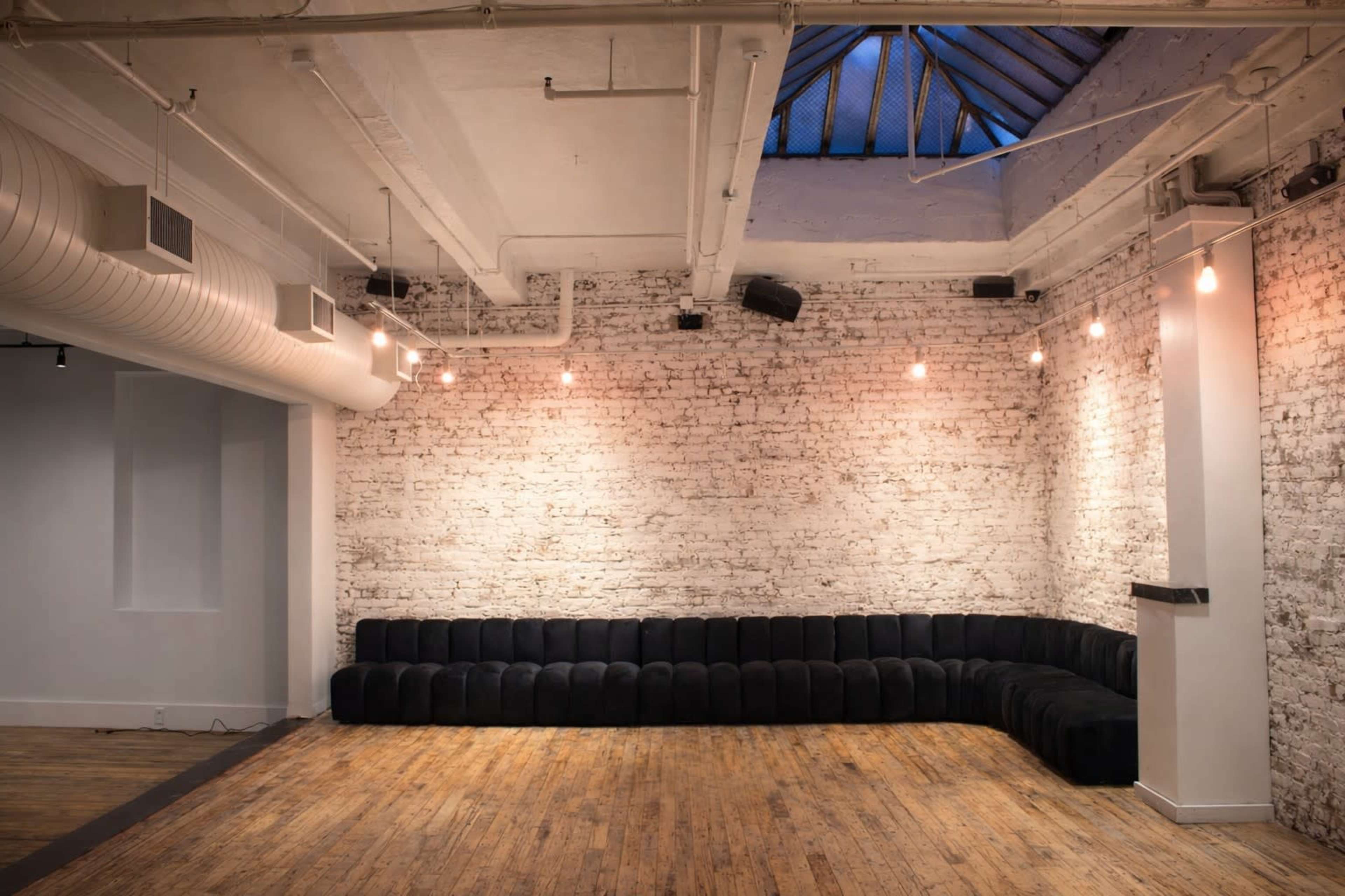 The image shows a spacious room with a brick wall and a large, curved black couch, illuminated by pendant lights hanging from the ceiling.