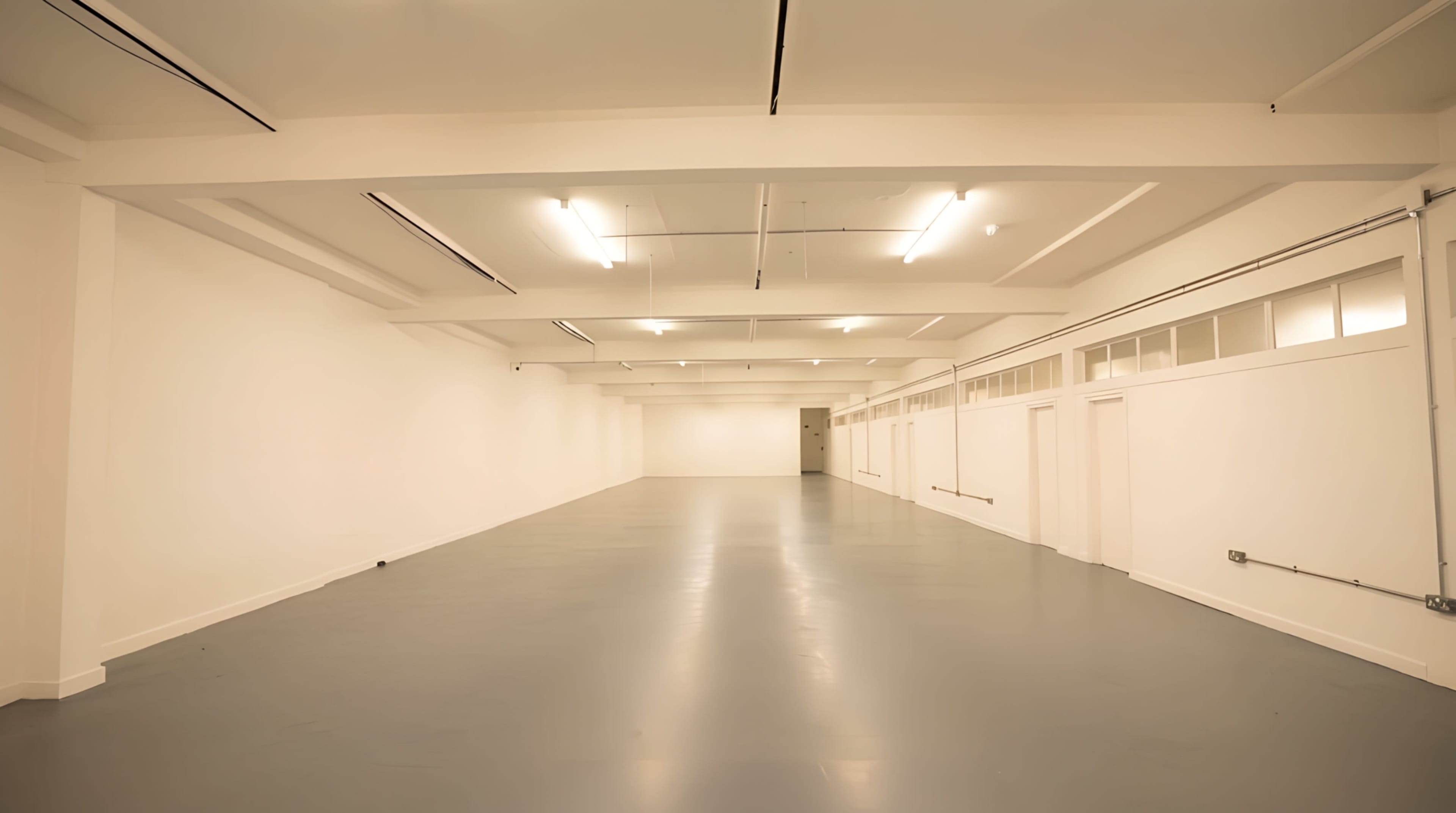 The image shows a large, empty room with white walls, a grey floor, and evenly spaced overhead lights.