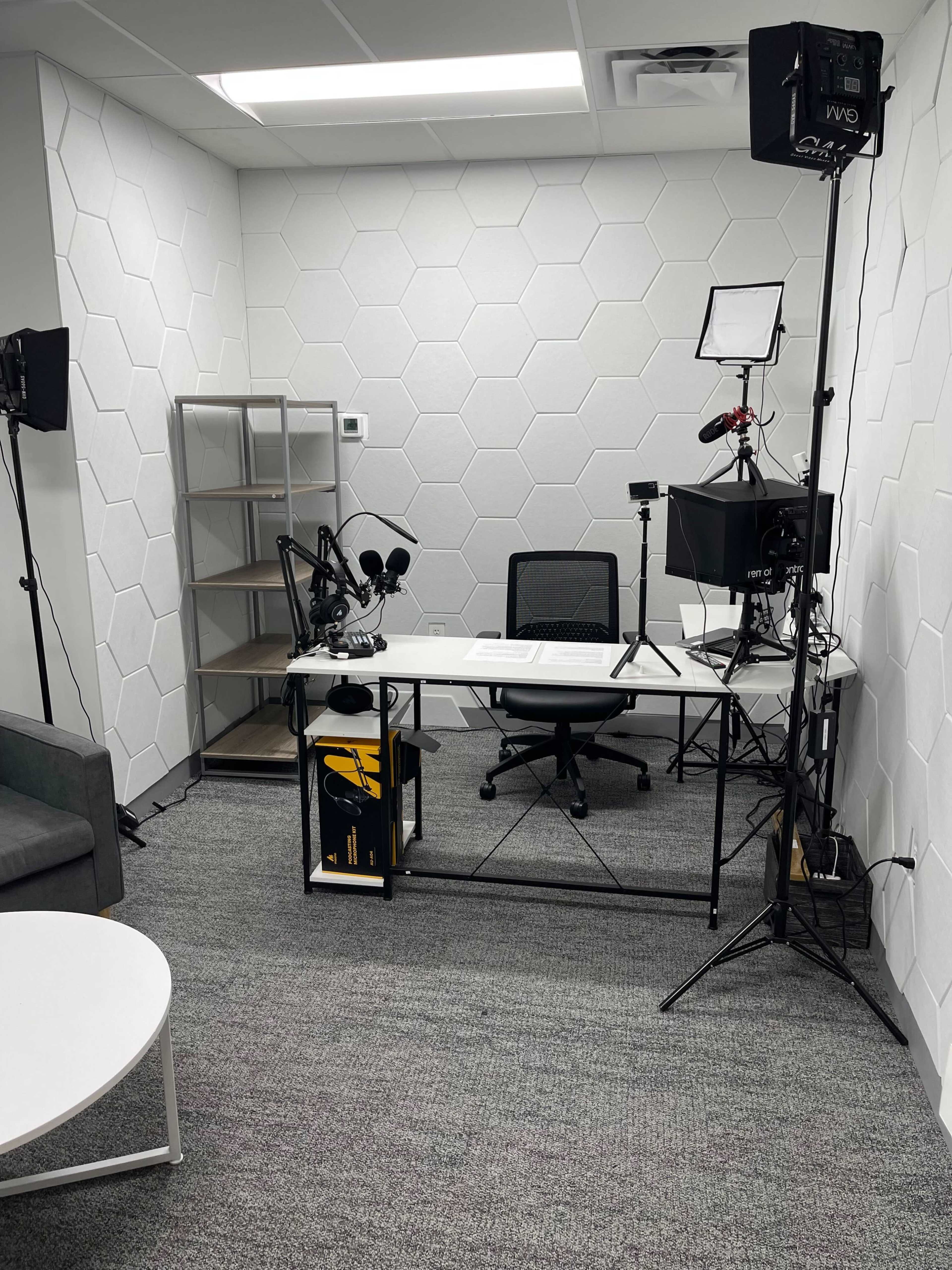 The image shows a modern recording studio setup with a desk, chairs, cameras, microphones, and a shelf in a room featuring hexagonal wall patterns.