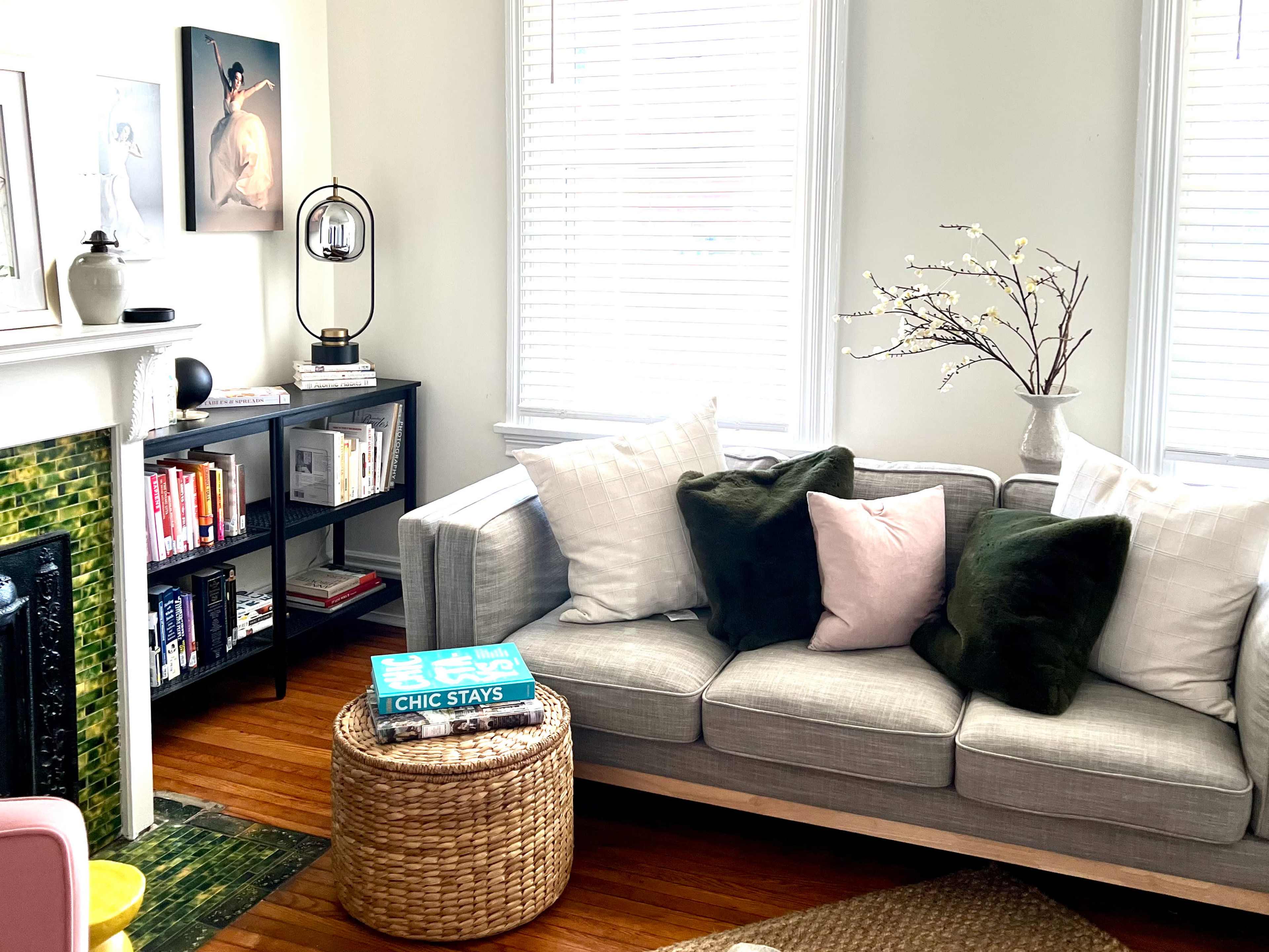 The image shows a cozy living room with a gray sofa adorned with various pillows, a wooden coffee table, and a bookshelf filled with books, alongside a decorative plant arrangement.