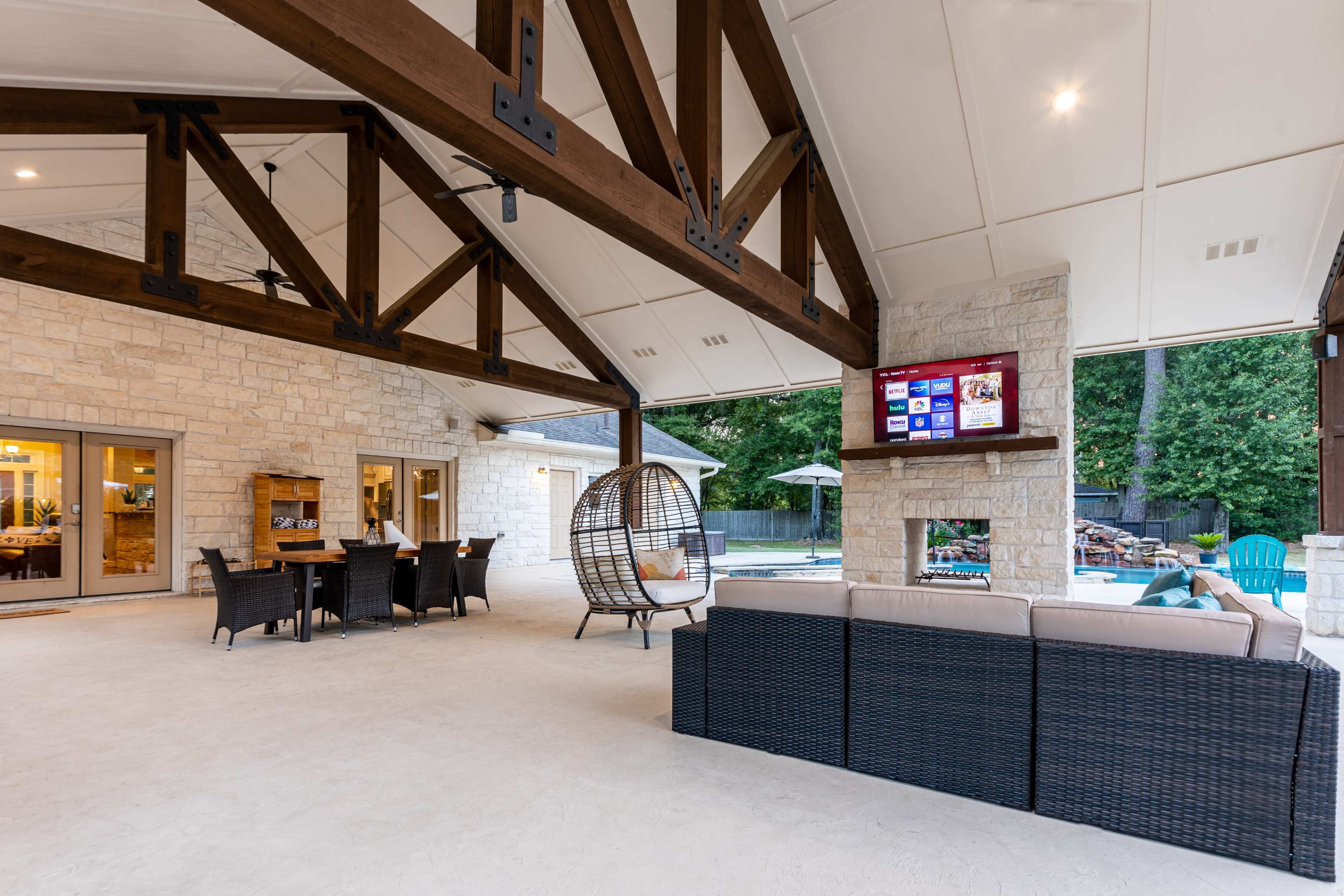 A covered patio area featuring a sectional sofa, a dining table, a hanging chair, and a television mounted above a stone fireplace, overlooking a swimming pool.