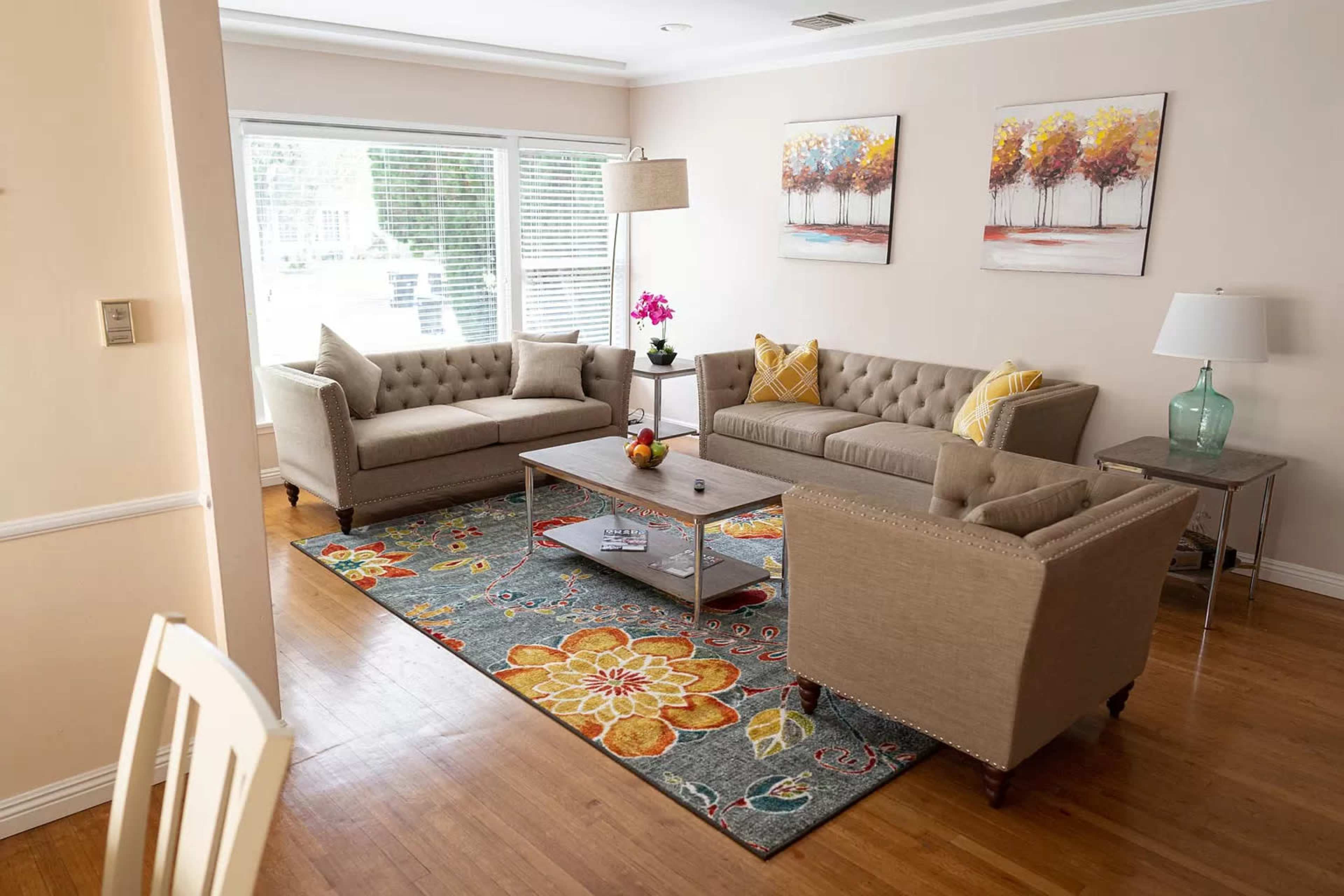 The image shows a living room with two tan sofas, a coffee table, and decorative artwork featuring trees on the walls, all arranged on a colorful area rug.
