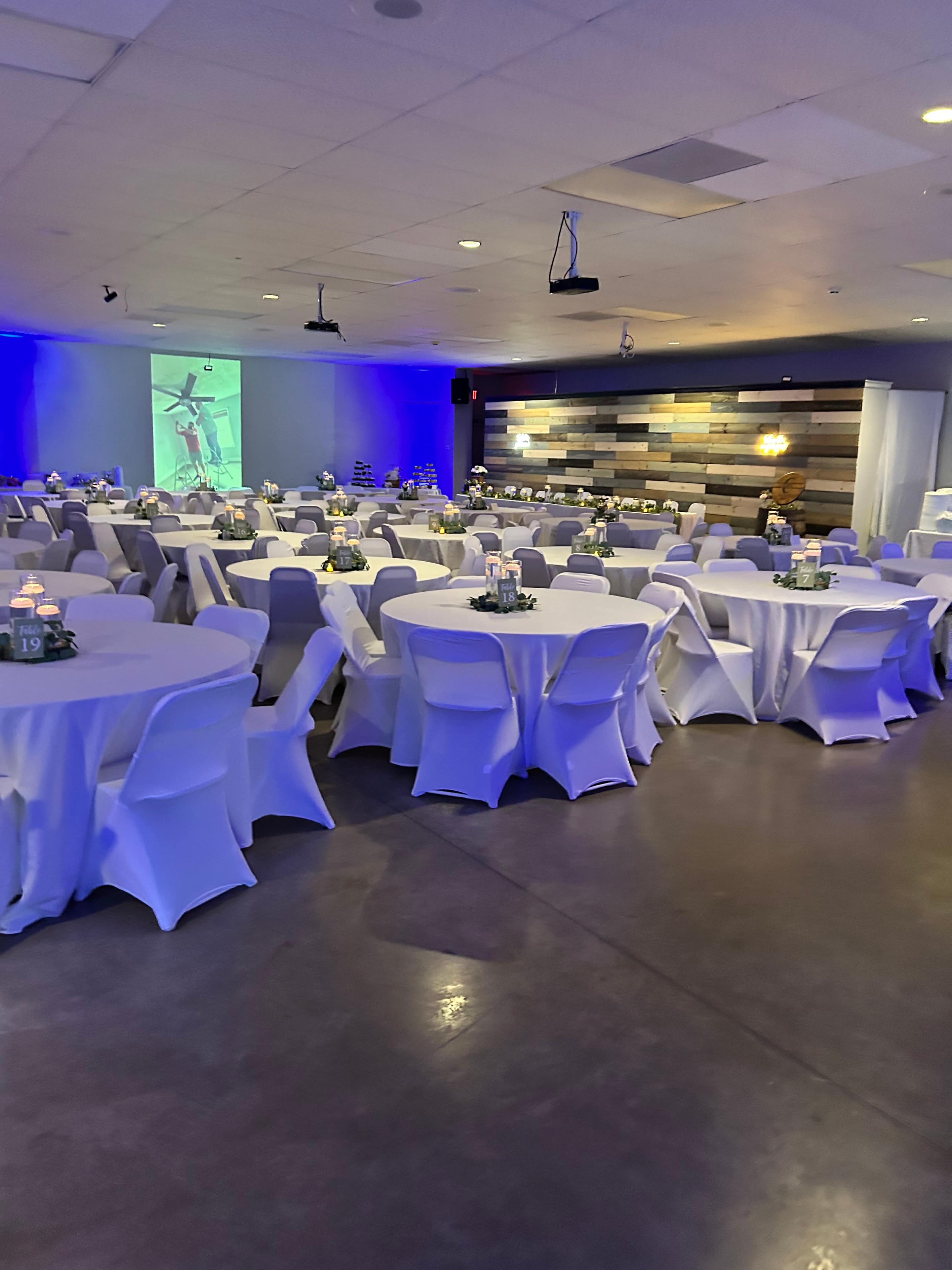 The image shows a spacious event room with round tables set with white tablecloths, and a soft blue light illuminating the area.